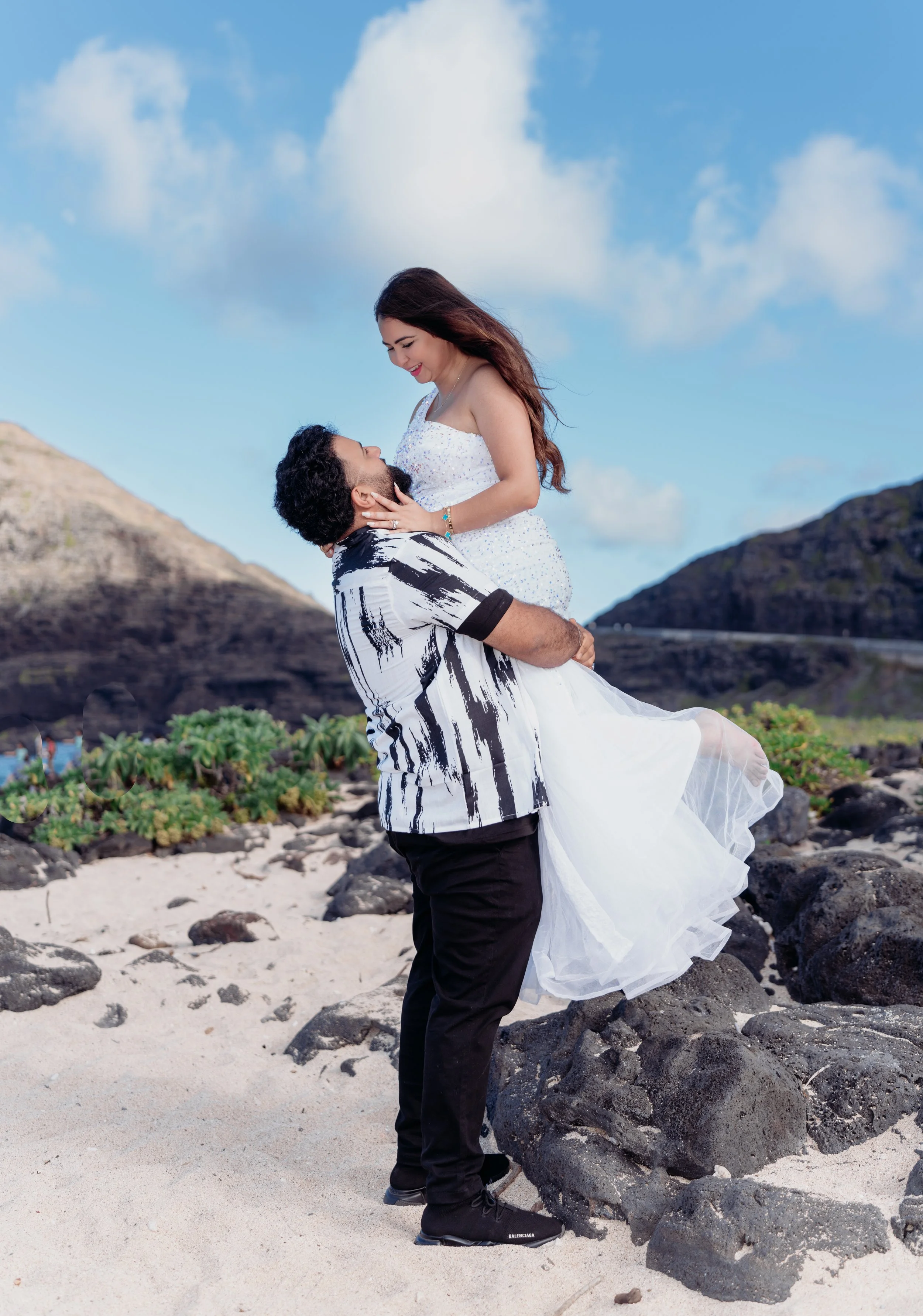 A man in a black and white patterned shirt lifts a woman in a white dress with blue accents on a rocky beach with sand and green plants, mountainous landscape in the background, under a partly cloudy sky.