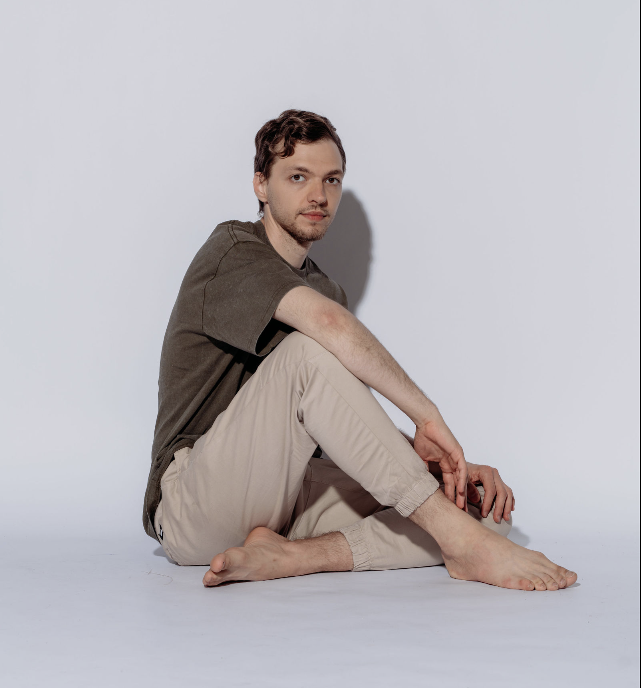 A young man with brown hair, wearing a dark T-shirt and beige pants, is sitting barefoot on a white surface against a plain white background, looking at the camera.
