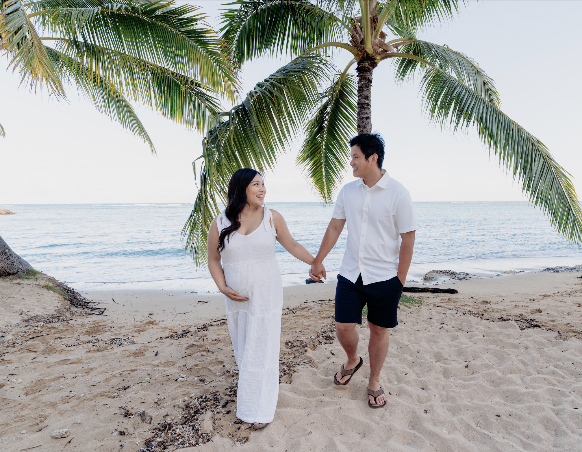 A happy couple walking hand in hand on a beach with palm trees, the woman appears pregnant, and they are smiling at each other.