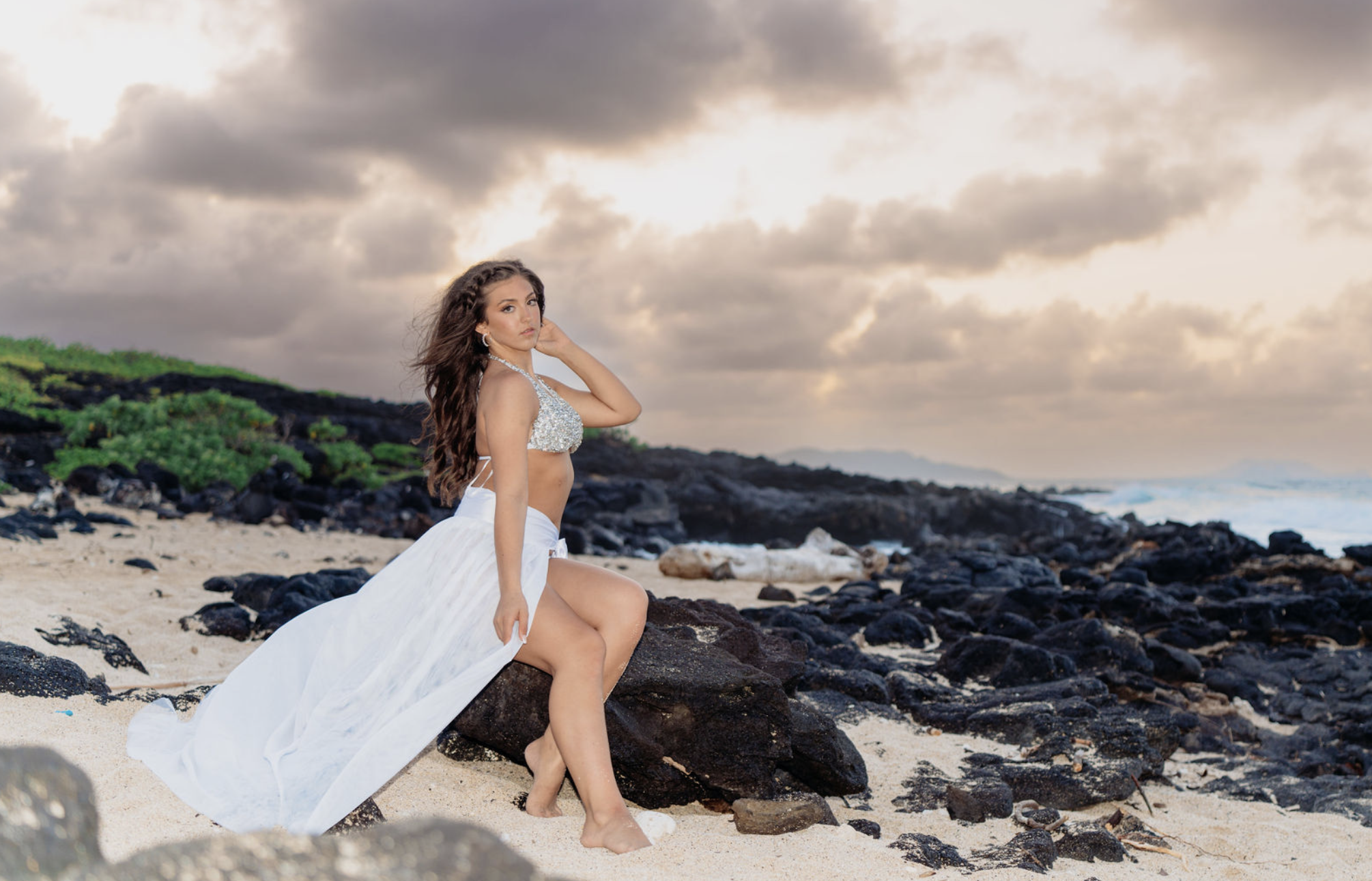 A woman in a white skirt and a sparkling crop top sits on a large rock on a sandy beach during sunset, with dark clouds and ocean waves in the background.
