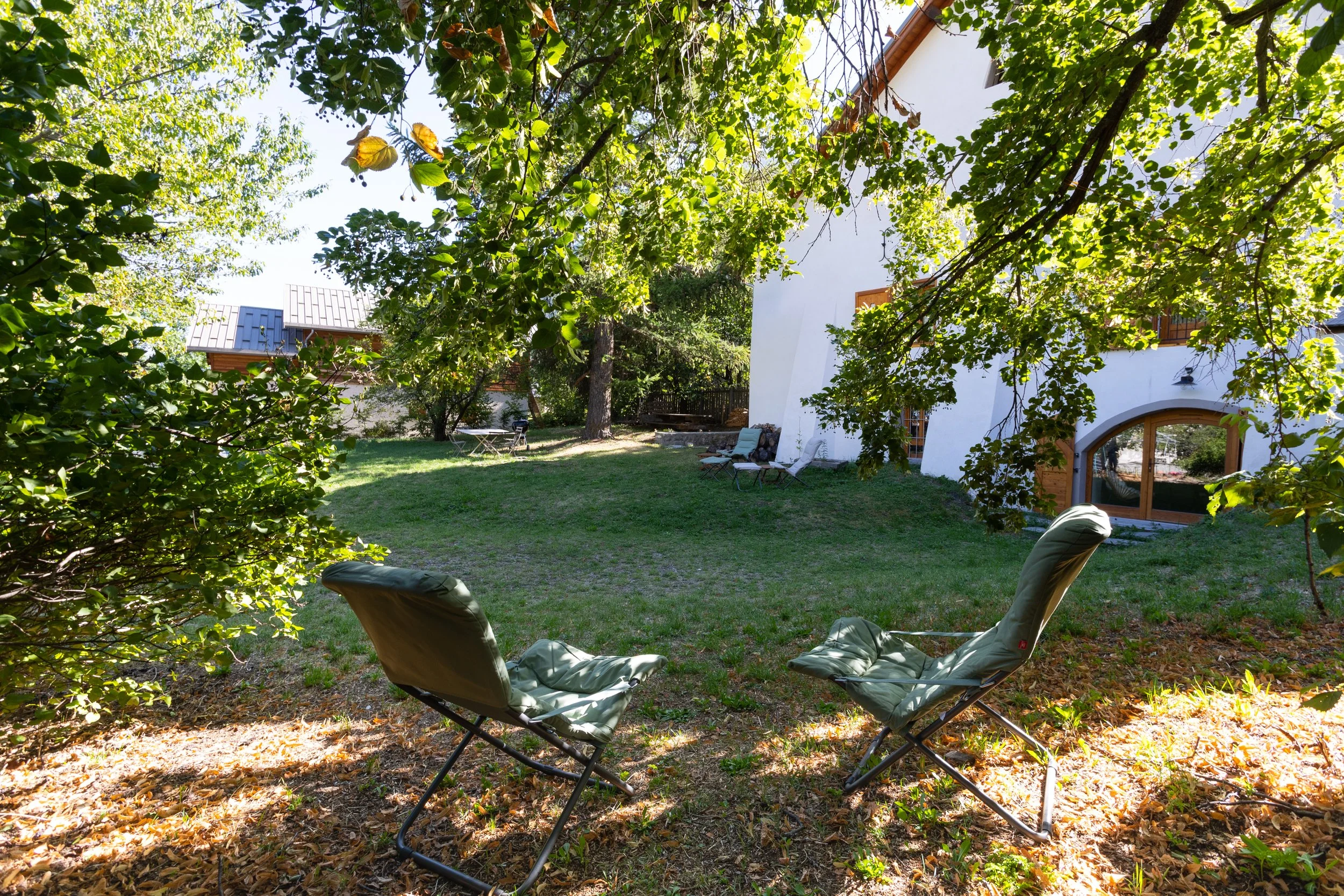 Deux fauteuils pliants sur un sol recouvert de feuilles mortes, sous un arbre, dans un jardin ensoleillé avec de l'herbe, et une maison blanche avec des fenêtres en bois à l'arrière-plan.