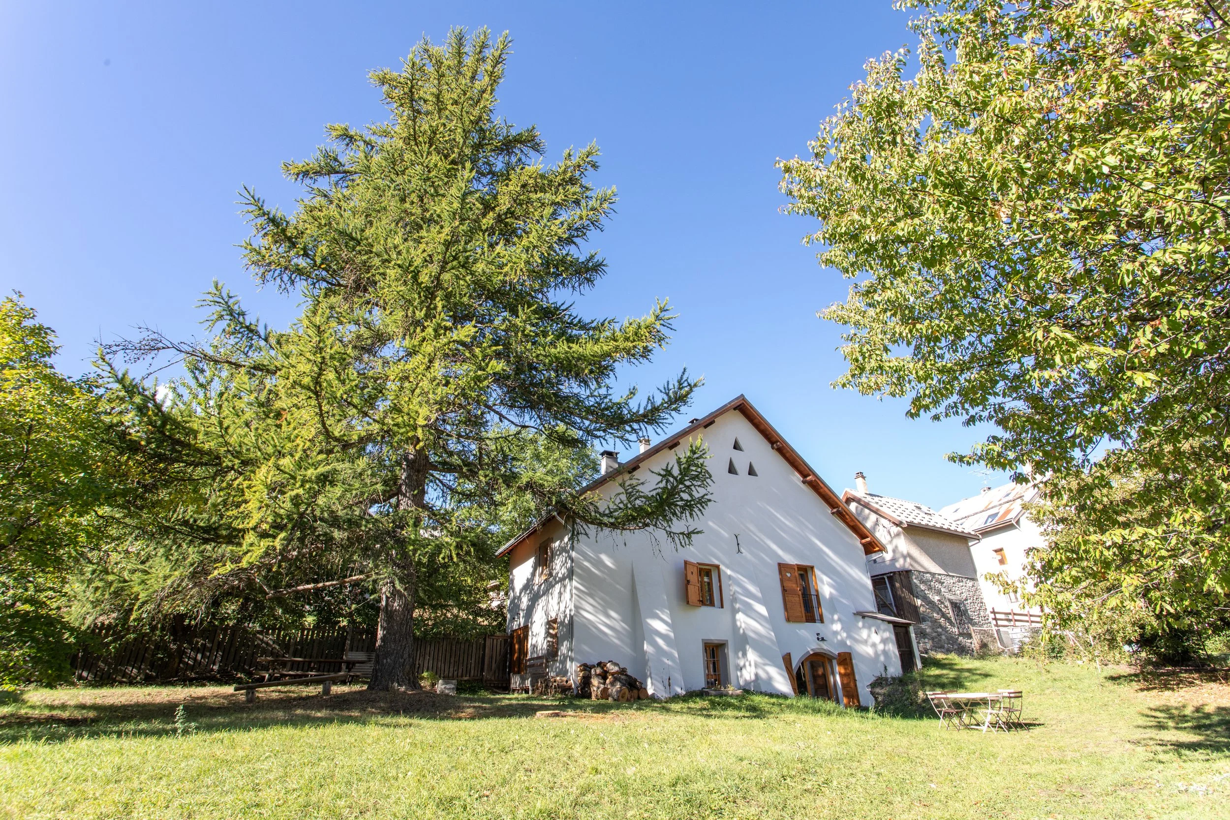 Maison blanche à toit en pente avec fenêtres en bois et volets, entourée d'arbres verts sous un ciel bleu clair, dans un jardin avec de l'herbe