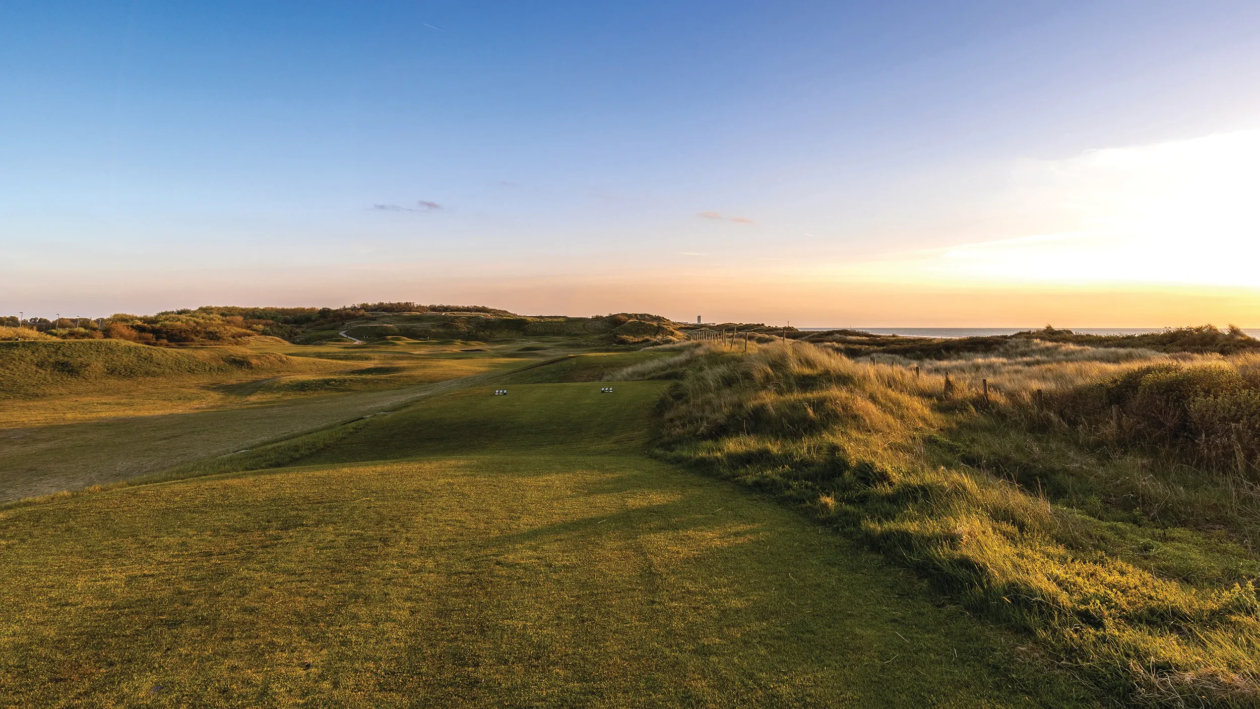Sunset over a golf course with a grassy fairway, sand dunes, and the ocean in the background.