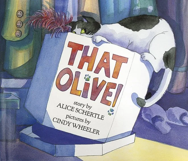A black and white cat hiding behind a large open book titled 'That Olive!' with a feathered toy near its head, in a cozy room with colorful curtains and a purple blanket.