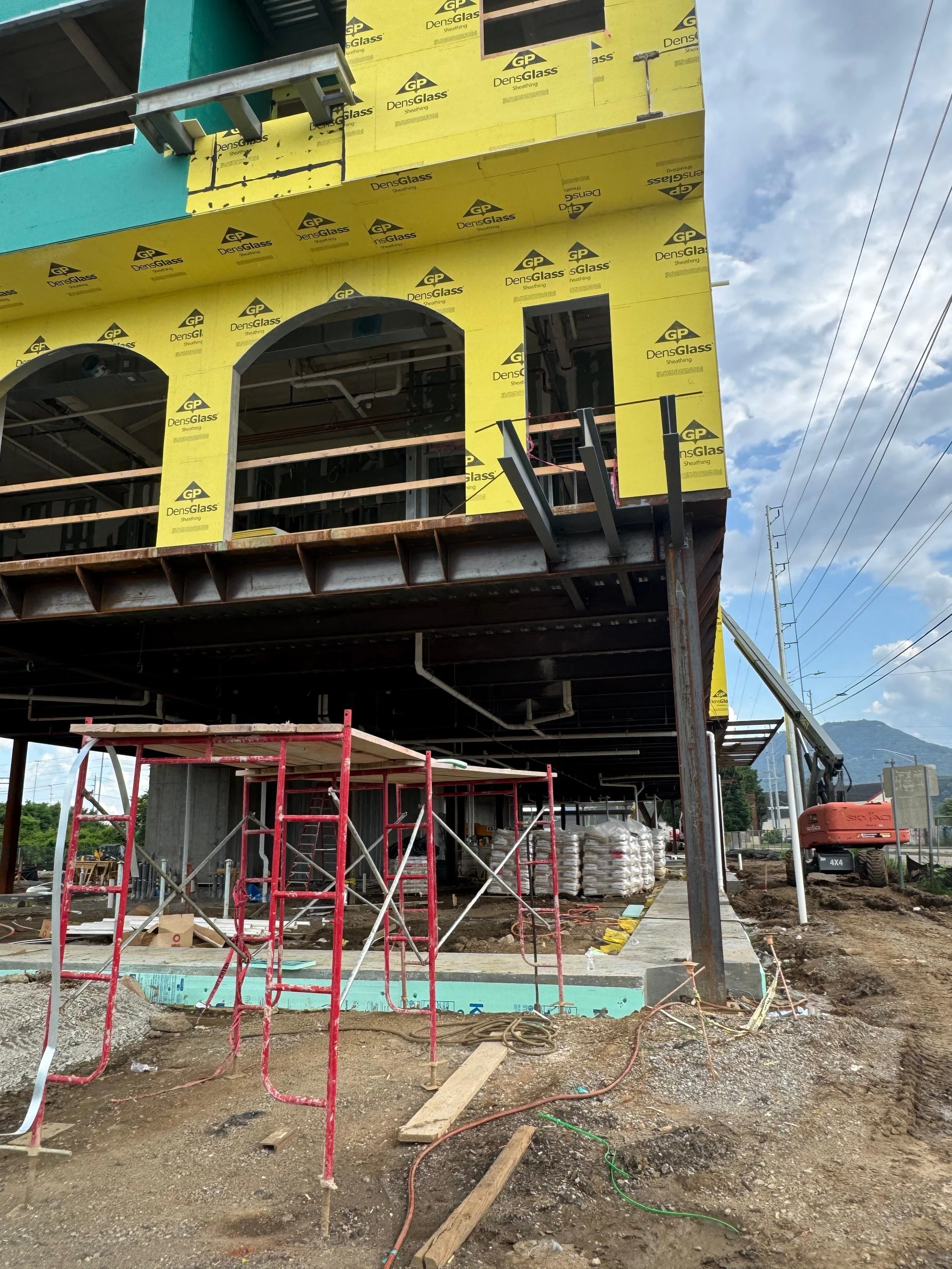 Construction site with a building under construction. The building has yellow insulation material and a turquoise wall, with scaffolding and construction equipment around.