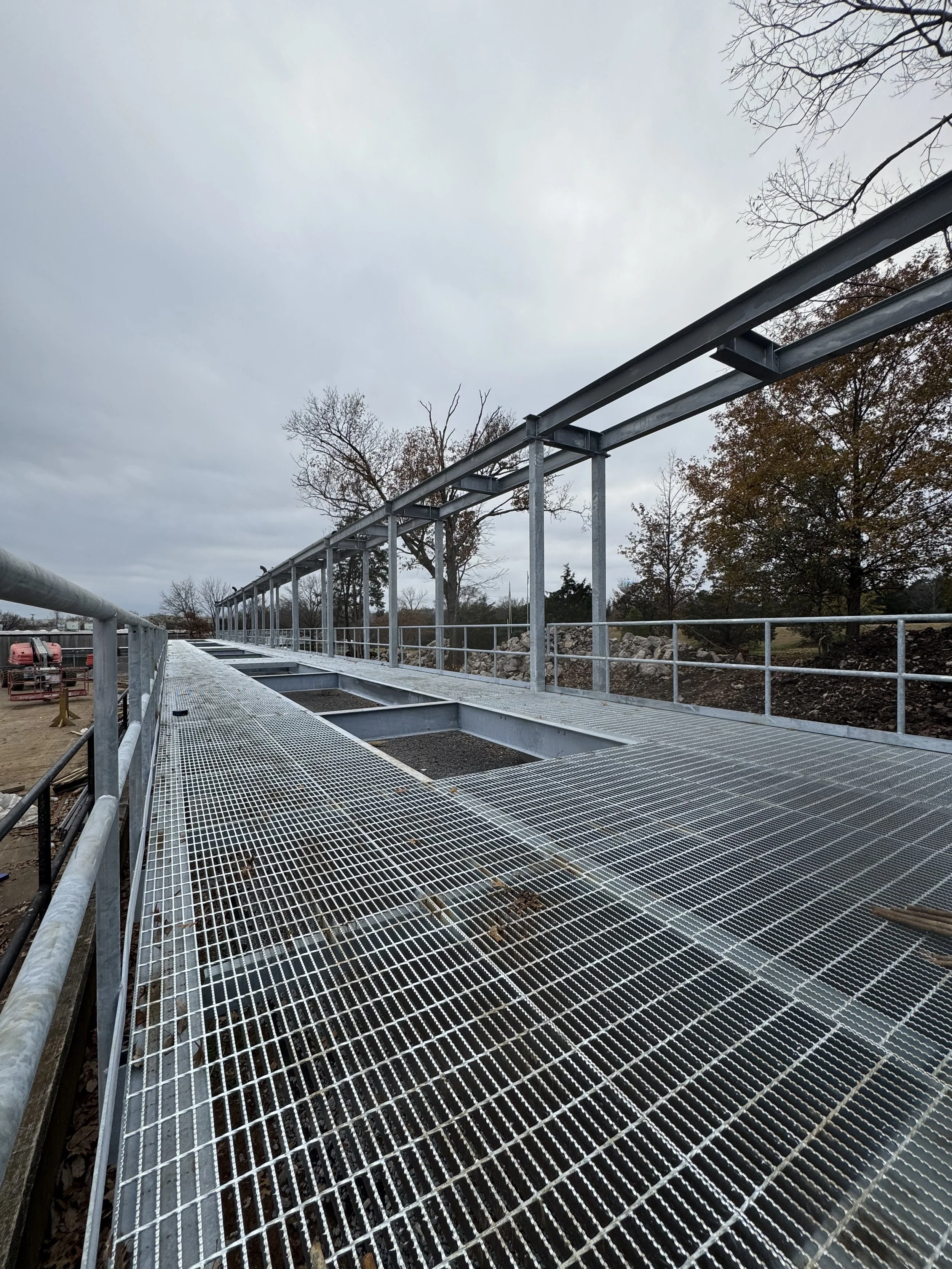 Construction site with metal scaffolding and grated platform under a cloudy sky, with trees in the background.