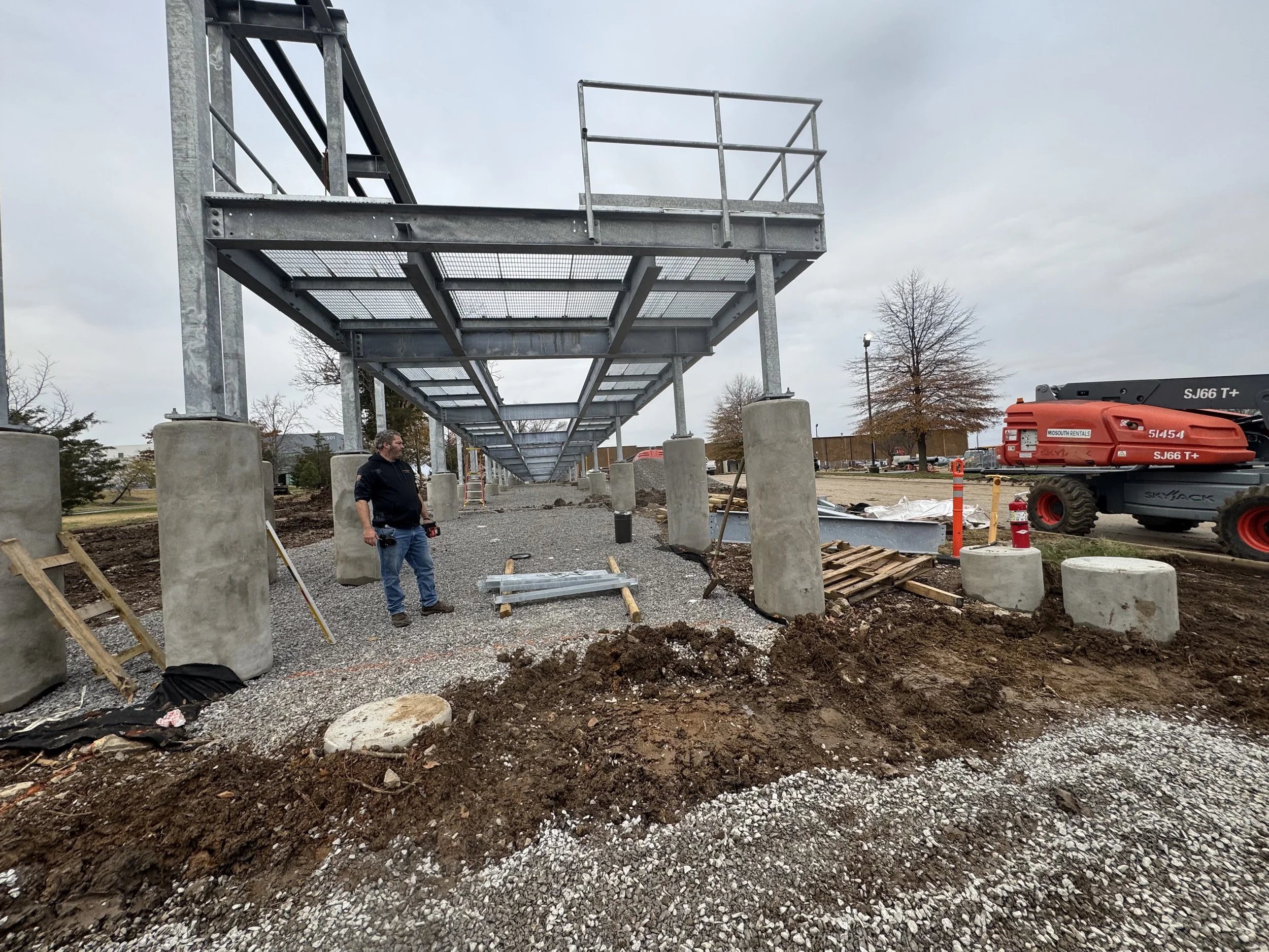 Construction site with steel structure, concrete pillars, excavation area, a worker with tools, and construction equipment under cloudy sky.