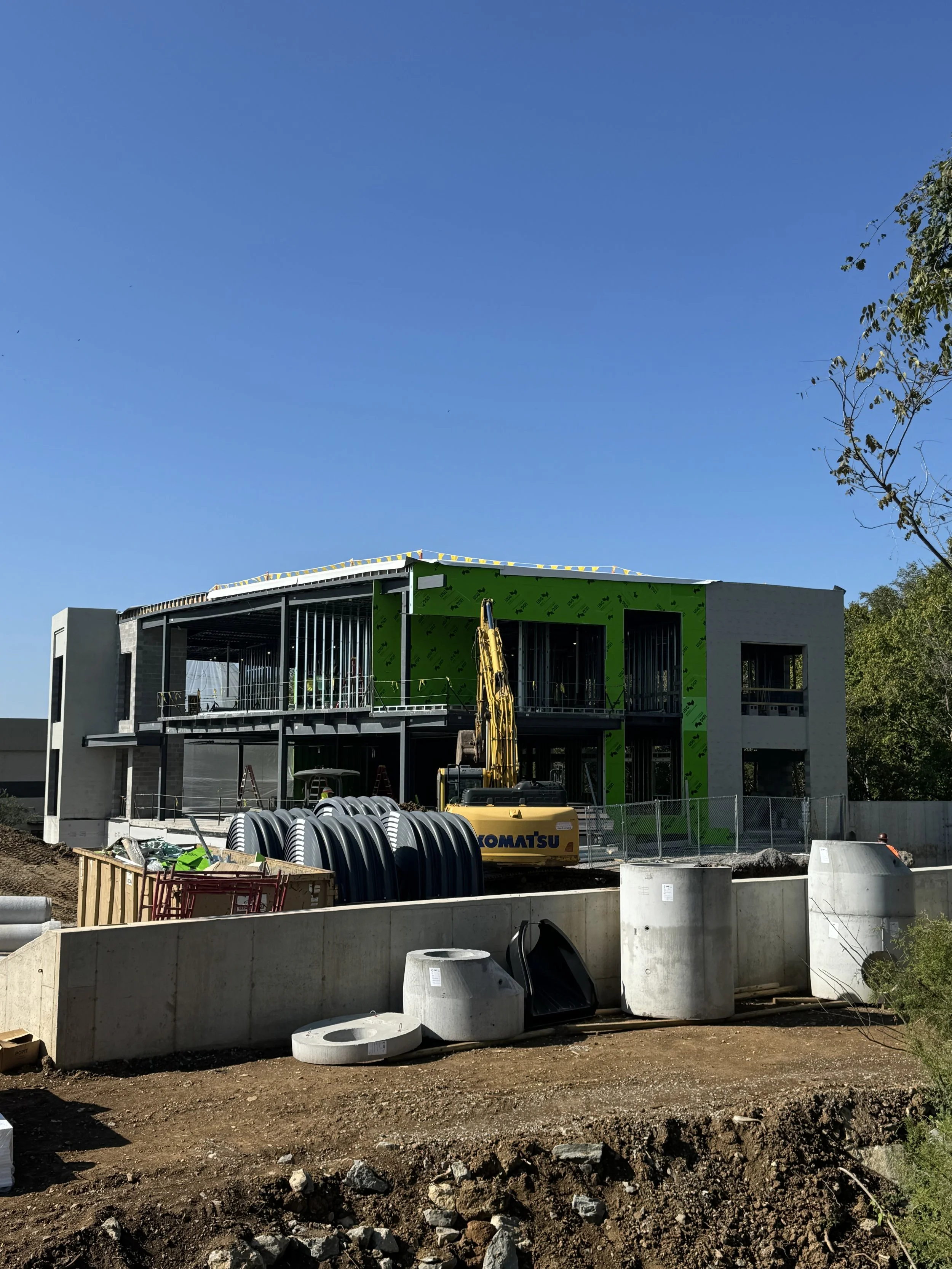 Construction site of a modern building with a yellow excavator, large concrete and plastic pipes, and green sheathing on part of the exterior walls. The building has an open upper balcony area and is surrounded by a chain-link fence.