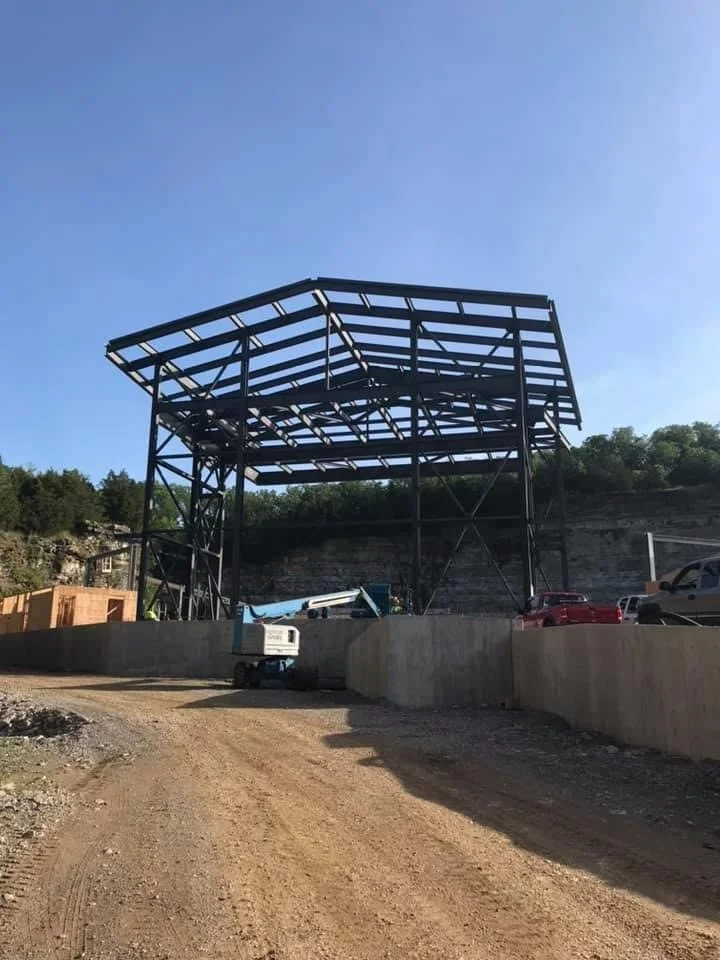 Steel framework structure being constructed on a construction site, with trucks and cars parked nearby, under a clear blue sky.