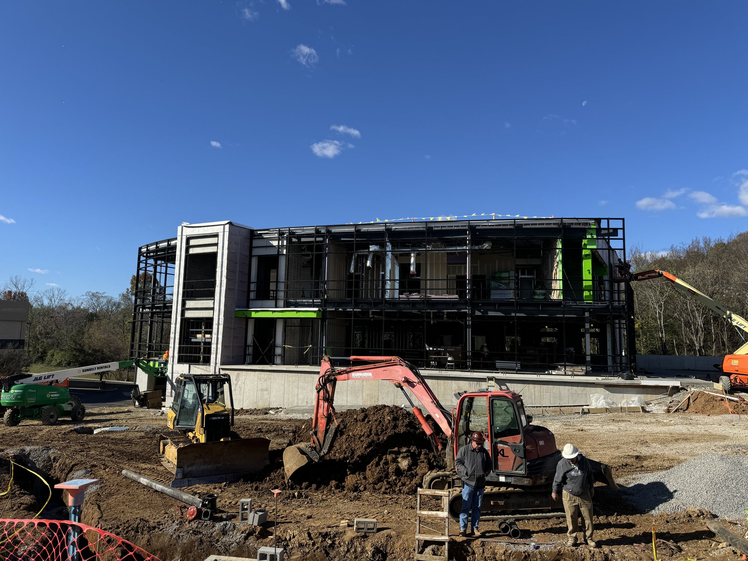 Construction site with a partially built multi-story building, heavy machinery like an excavator, and construction workers under a clear blue sky.