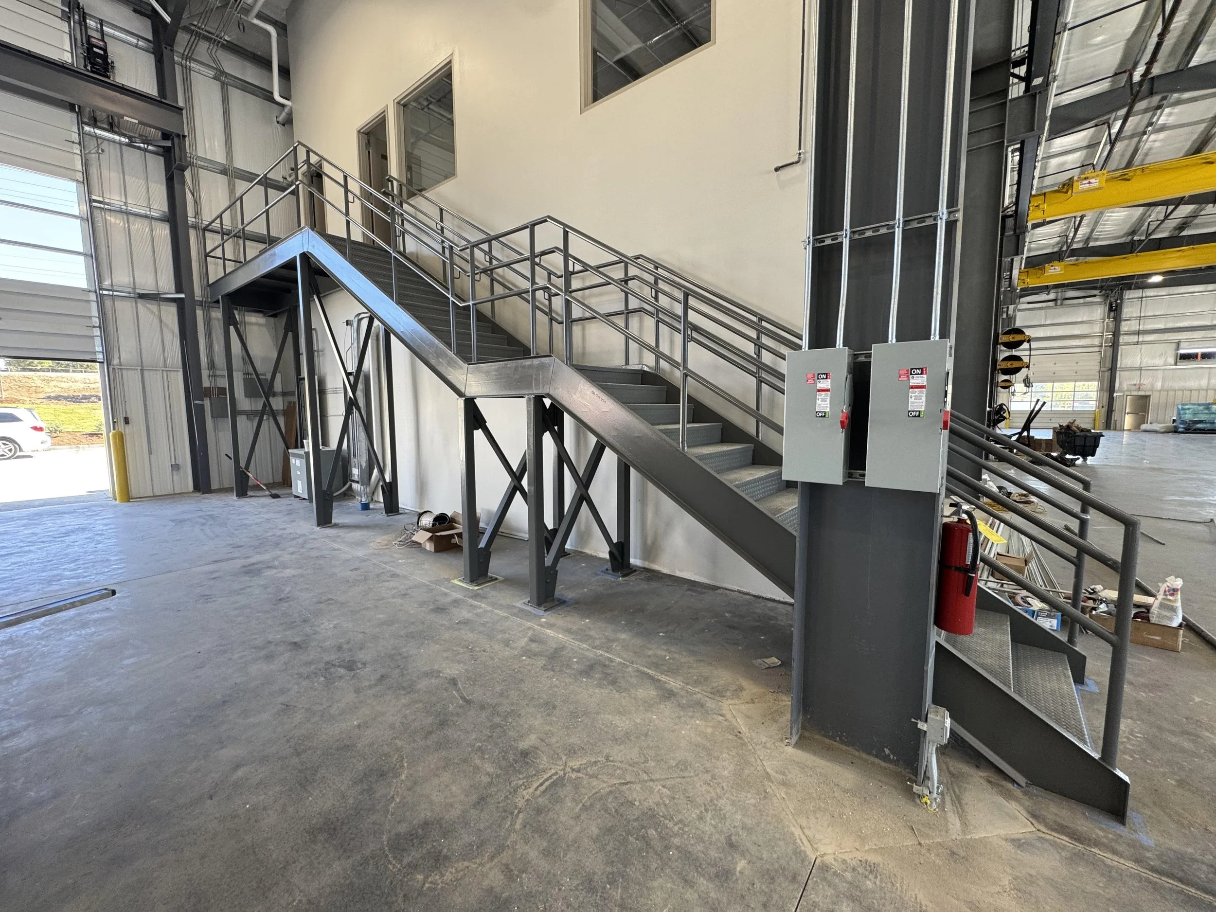 Industrial staircase with metal handrails leading to a second-floor entrance in a warehouse or factory setting, with electrical panels mounted on the side of the stairs and safety equipment nearby.