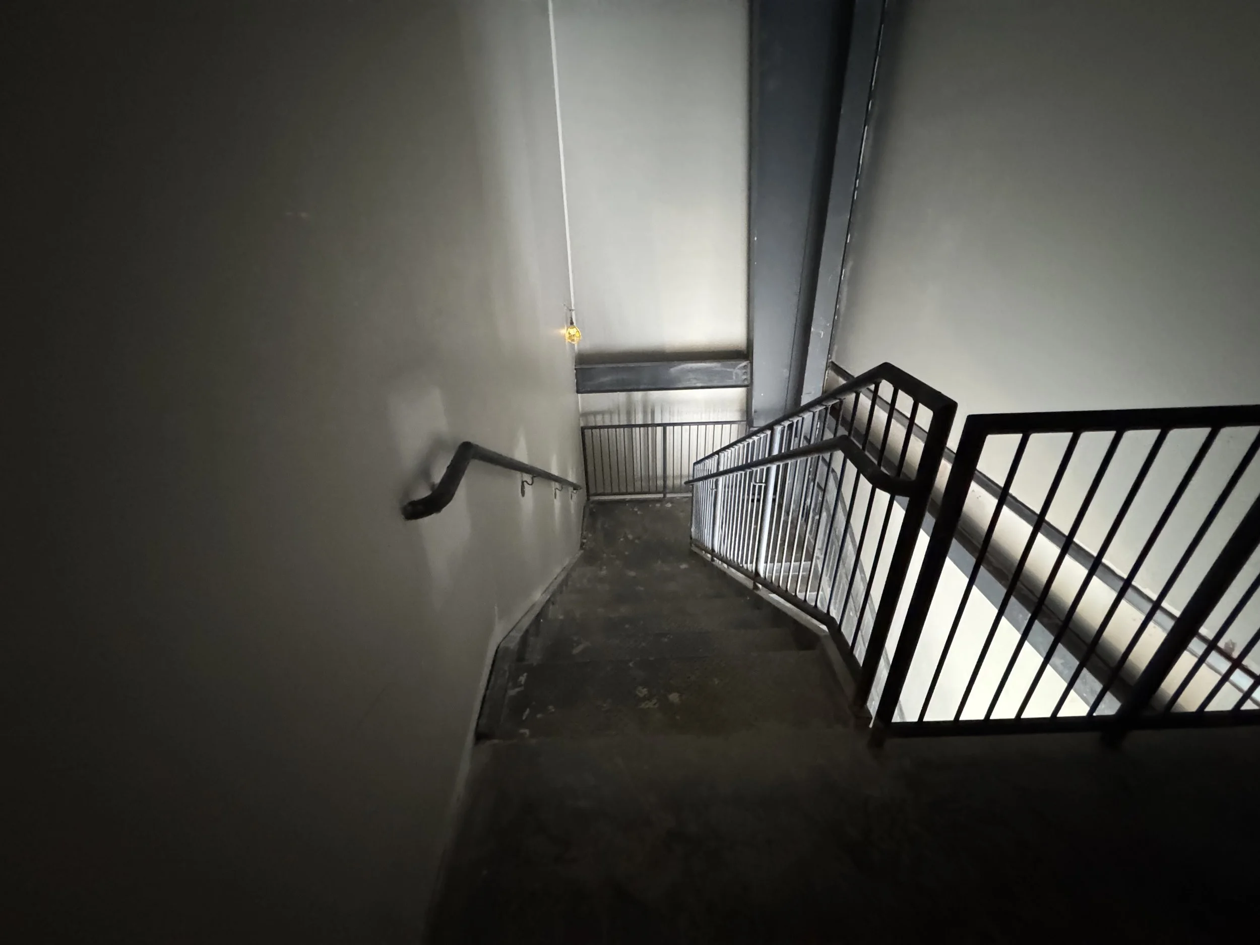 Dark stairwell with metal railing and a smooth wall, illuminated by dim light at the top, leading down to a landing and another staircase.