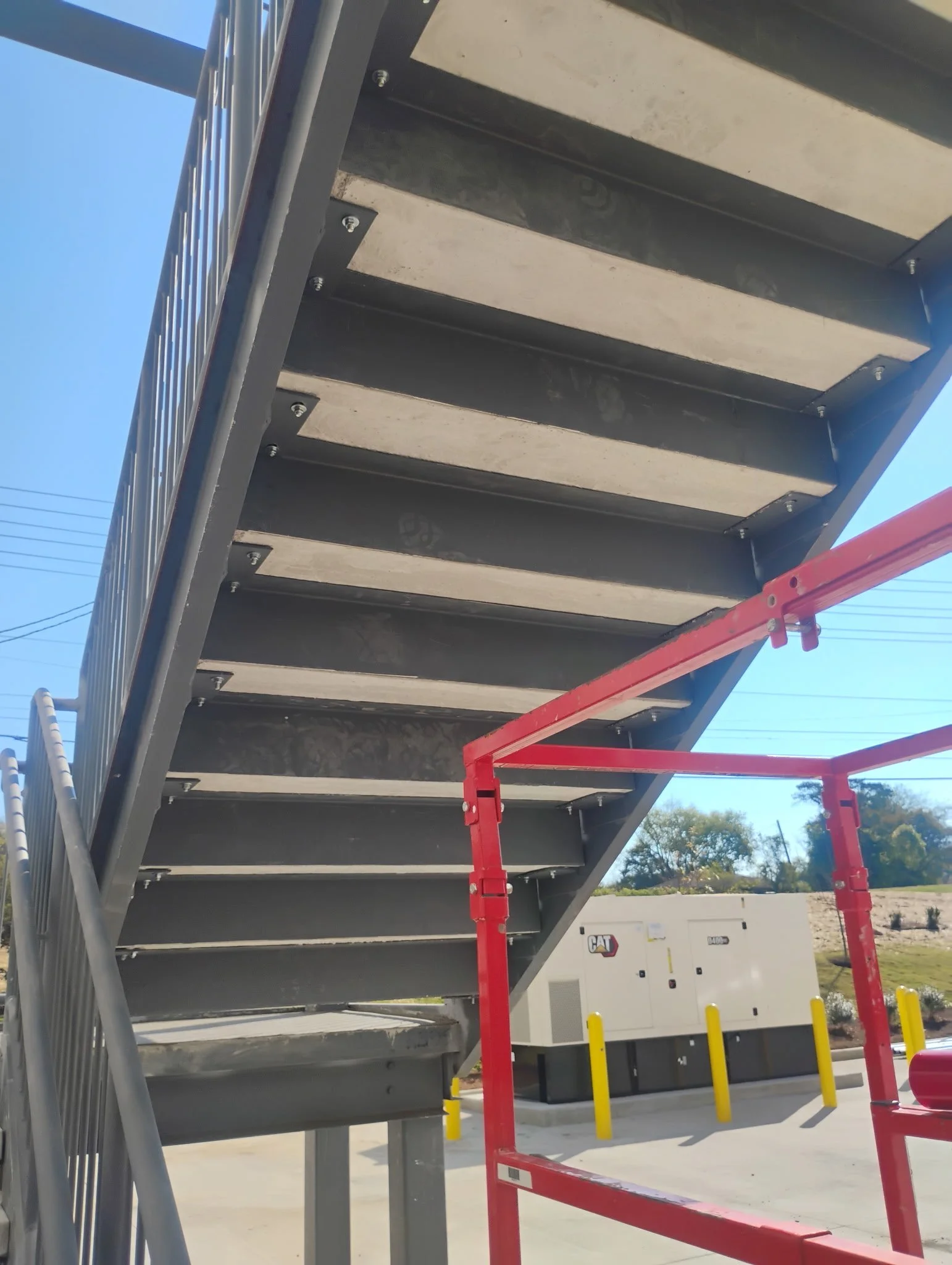 Underneath view of metal staircase with black steps and silver railings, red scaffolding, and a white generator in the background with the CAT logo.