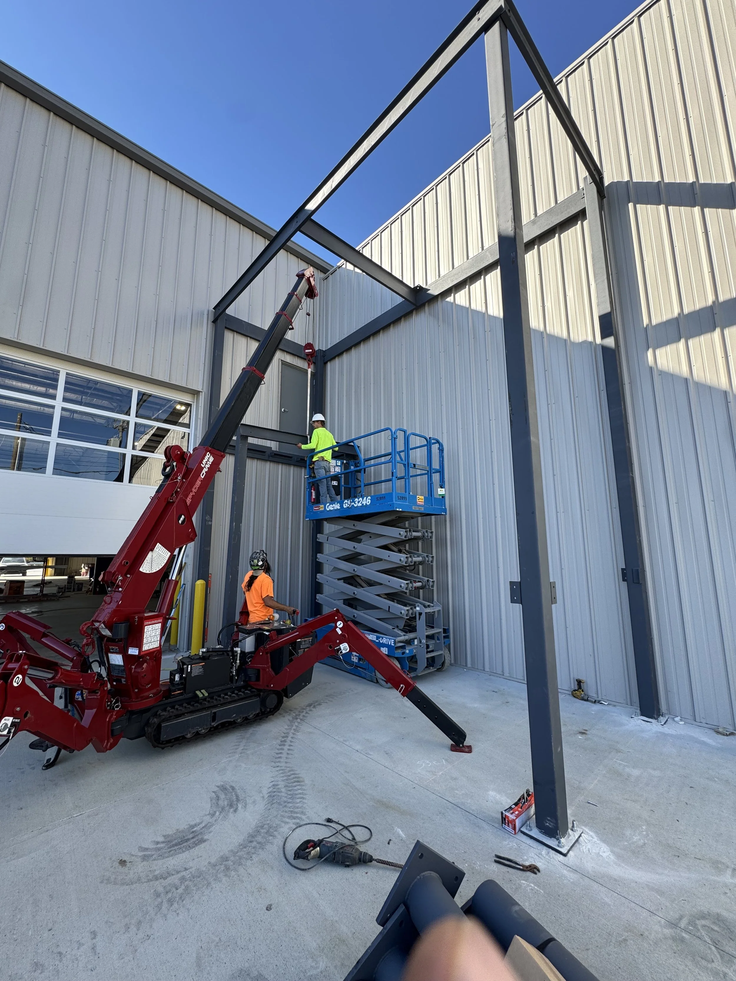 Construction site with a red crane and two workers, one in a neon yellow shirt and another in an orange shirt, assembling a steel structure outside a warehouse.