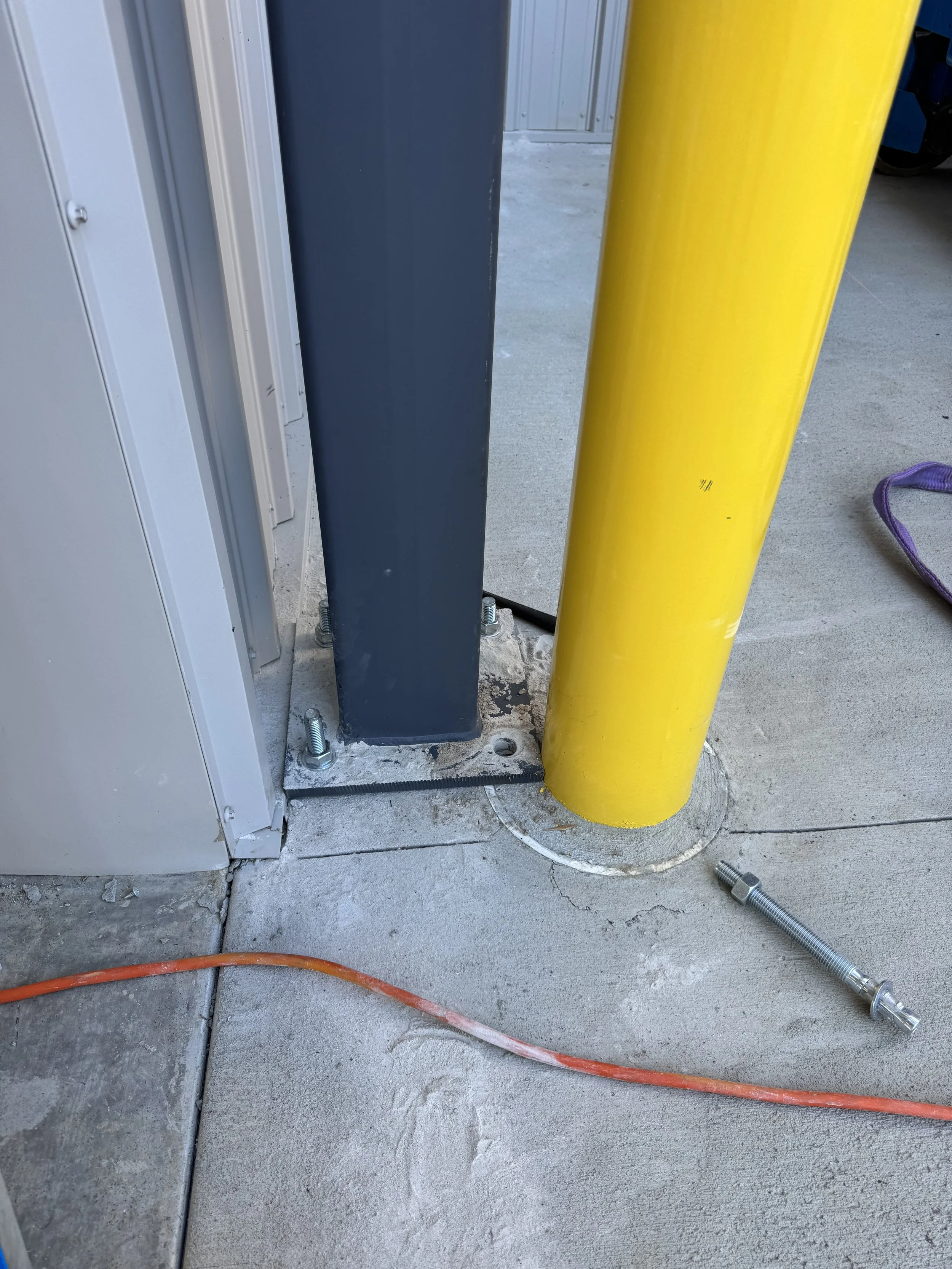 Close-up of a construction site showing a yellow safety pole, a gray metal post, a gray electrical box, and an orange extension cord on a concrete floor.