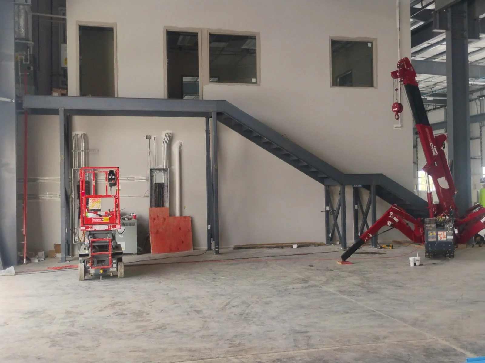 Construction site with a red crane and a metal staircase leading to a second-floor office space with three windows.