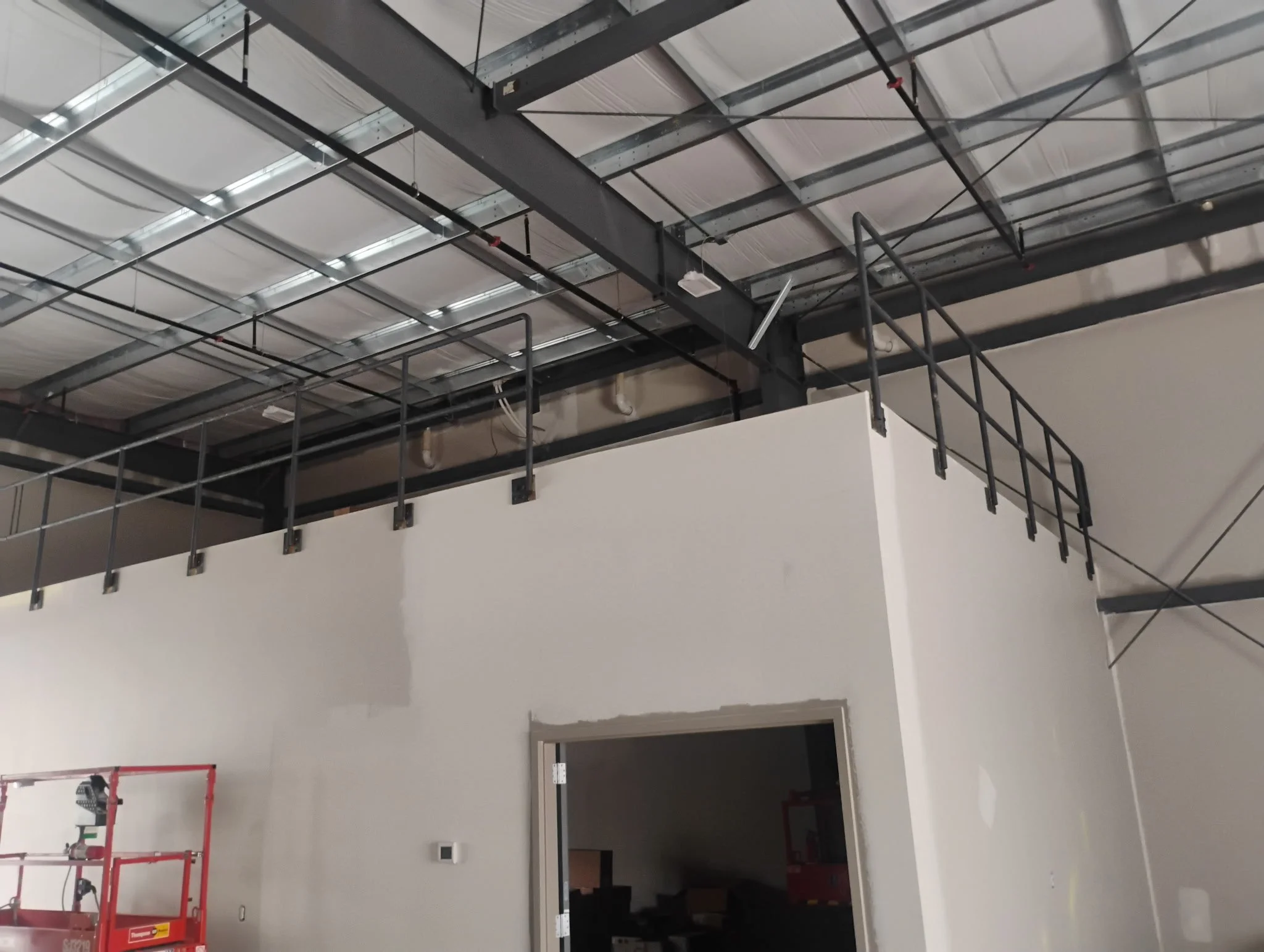 Construction site interior showing a second-floor metal balcony with guardrails, an unfinished wall, and an open doorway, with construction equipment and supplies around.