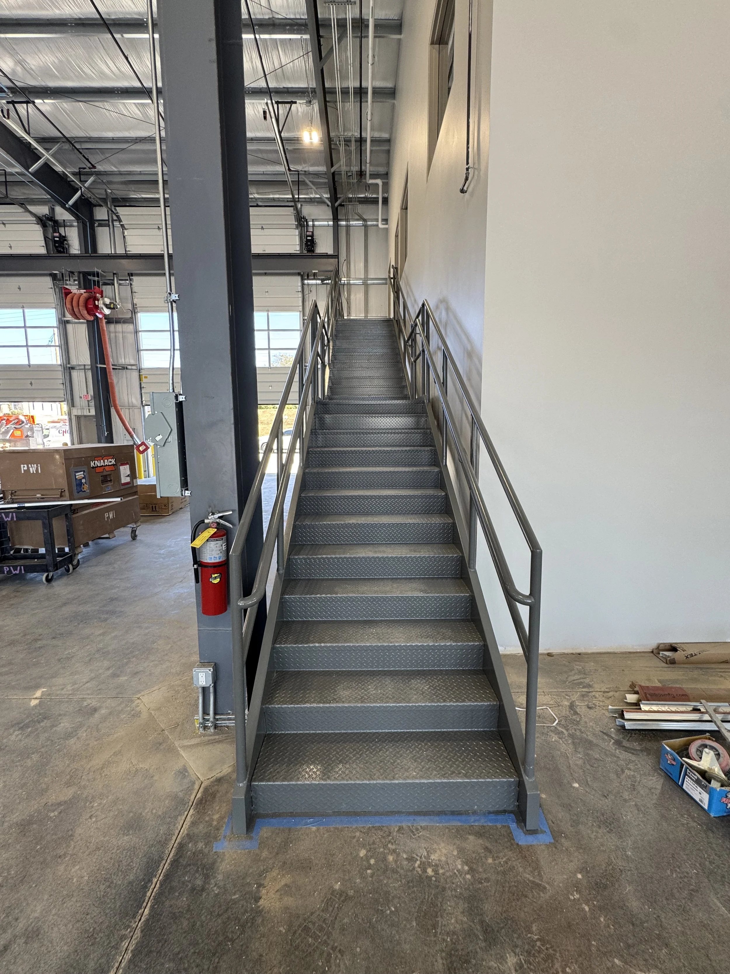 Metal stairway inside a warehouse or industrial building, with handrails on both sides and a concrete floor.