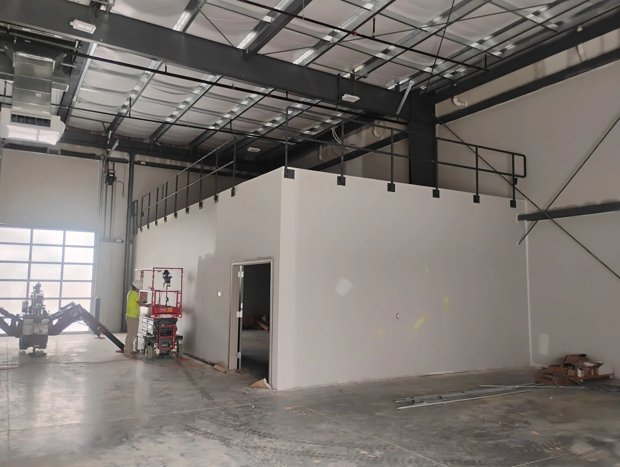 Interior of a building under construction with workers and scaffolding, and a wall with a door. The ceiling has exposed beams and insulation.
