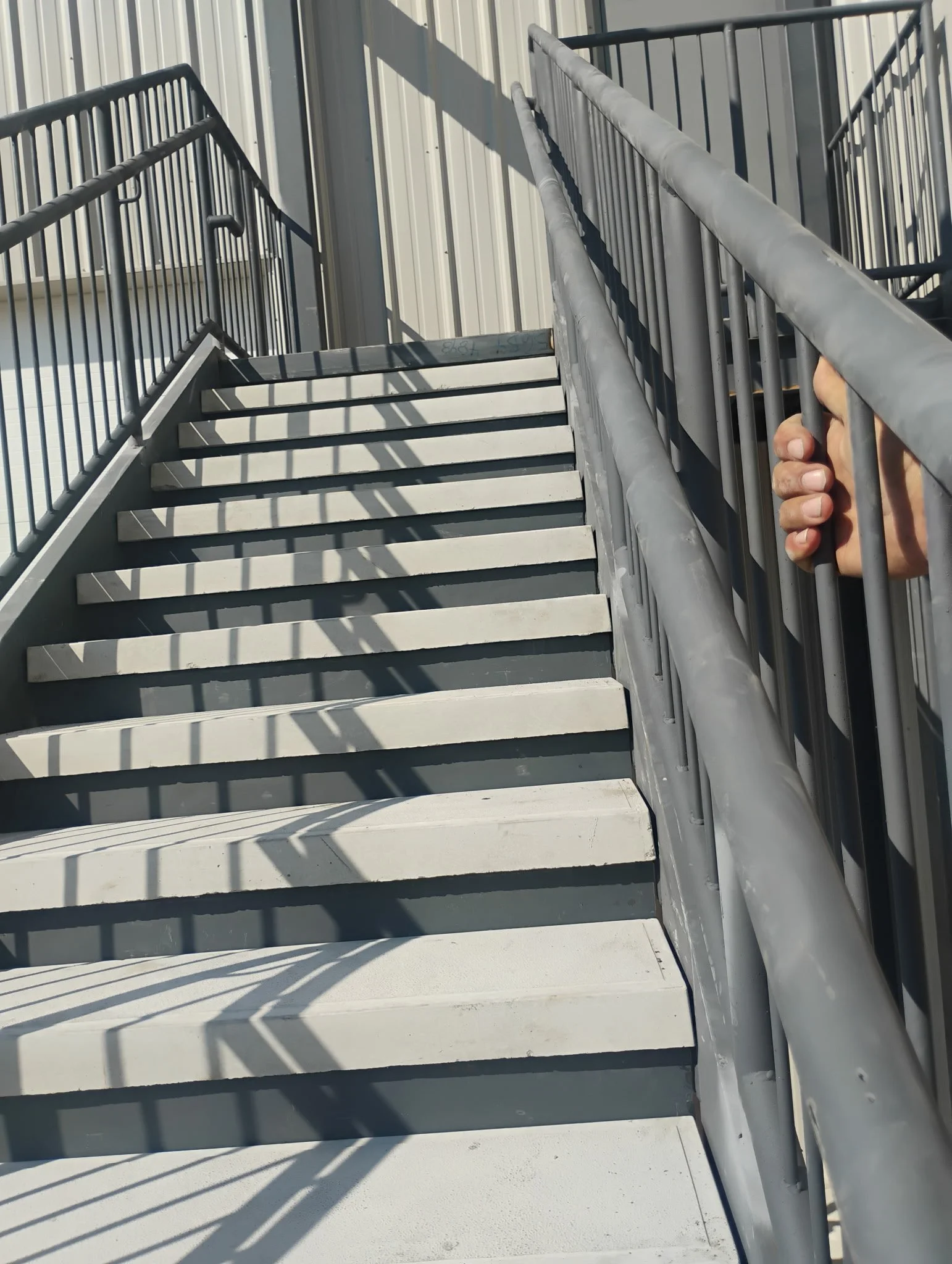 Person holding metal railing on outdoor metal staircase with shadow patterns cast on the steps.