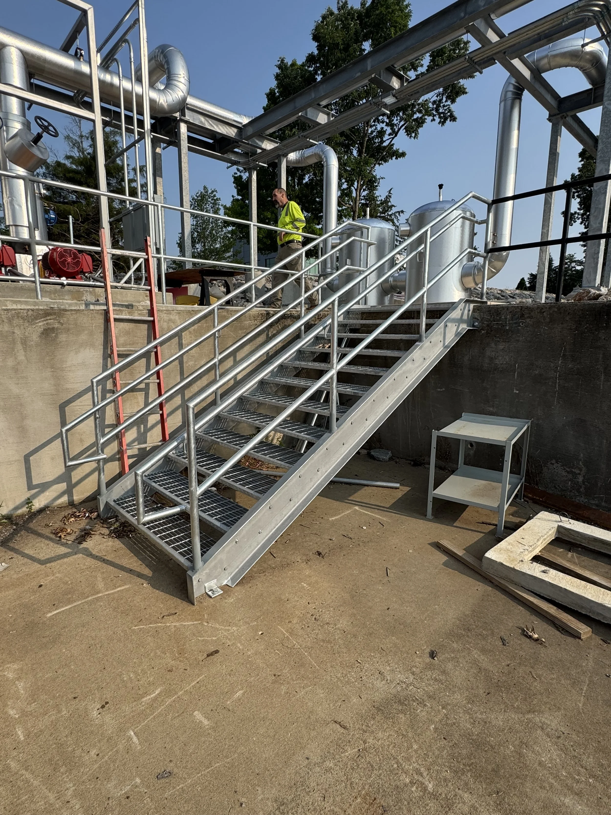 Metal staircase leading up to an industrial piping system at a construction site with a worker in a yellow safety vest.