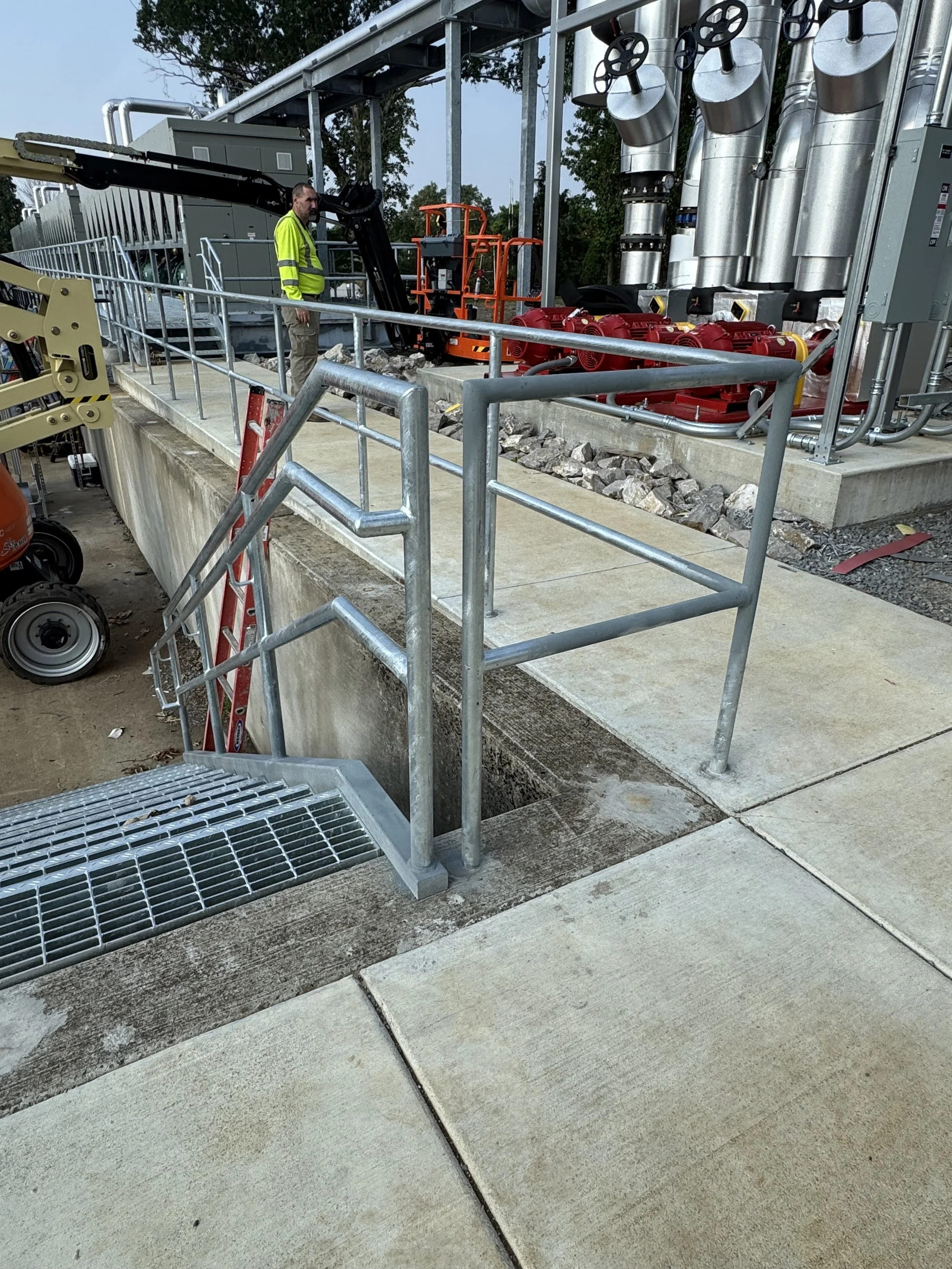 Construction site with metal railings, stairs, and equipment, including a worker in a safety vest and machinery in the background.