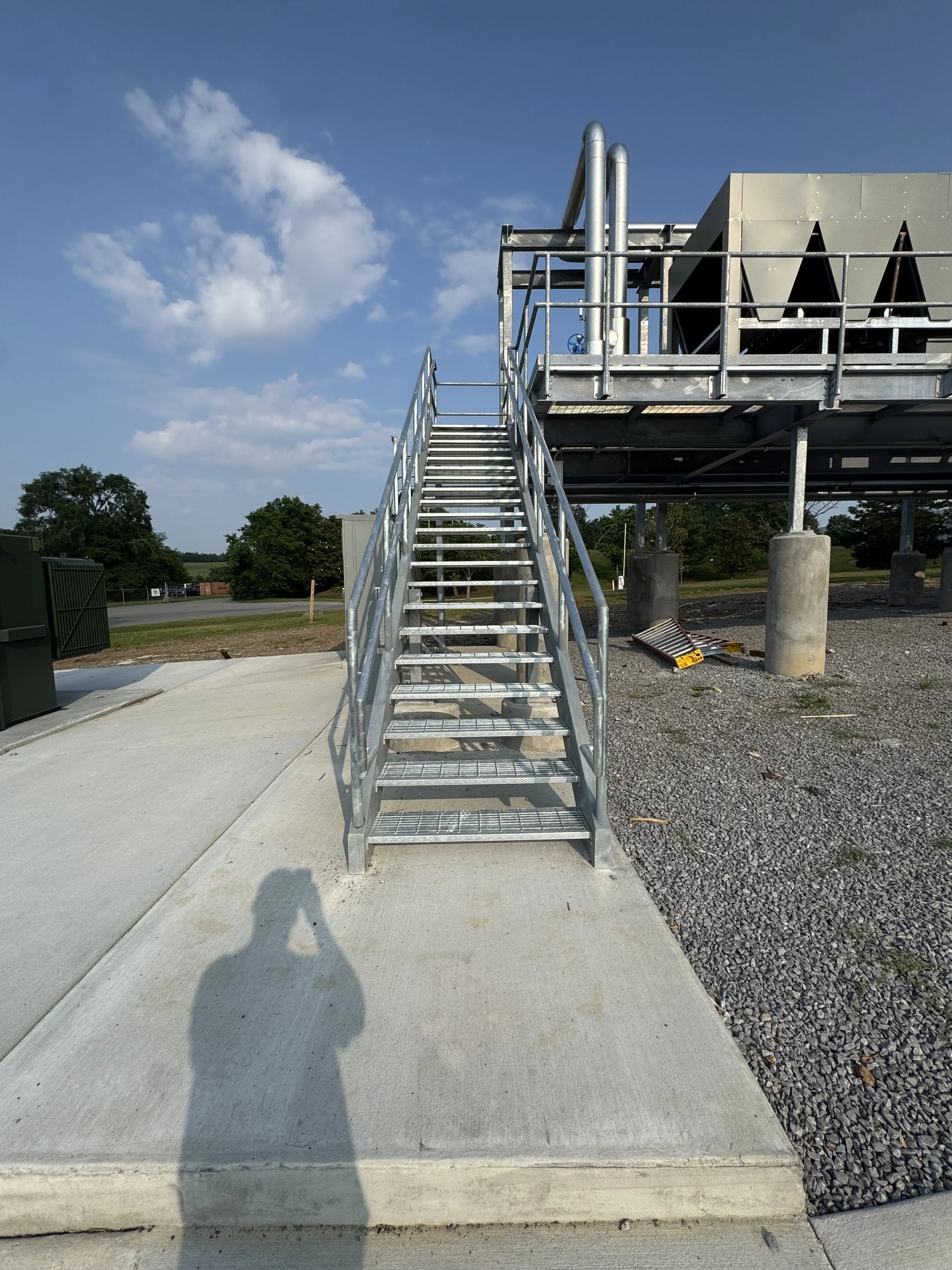 Metal staircase leading up to a raised industrial platform with safety railings, set on concrete and gravel under a partly cloudy sky.