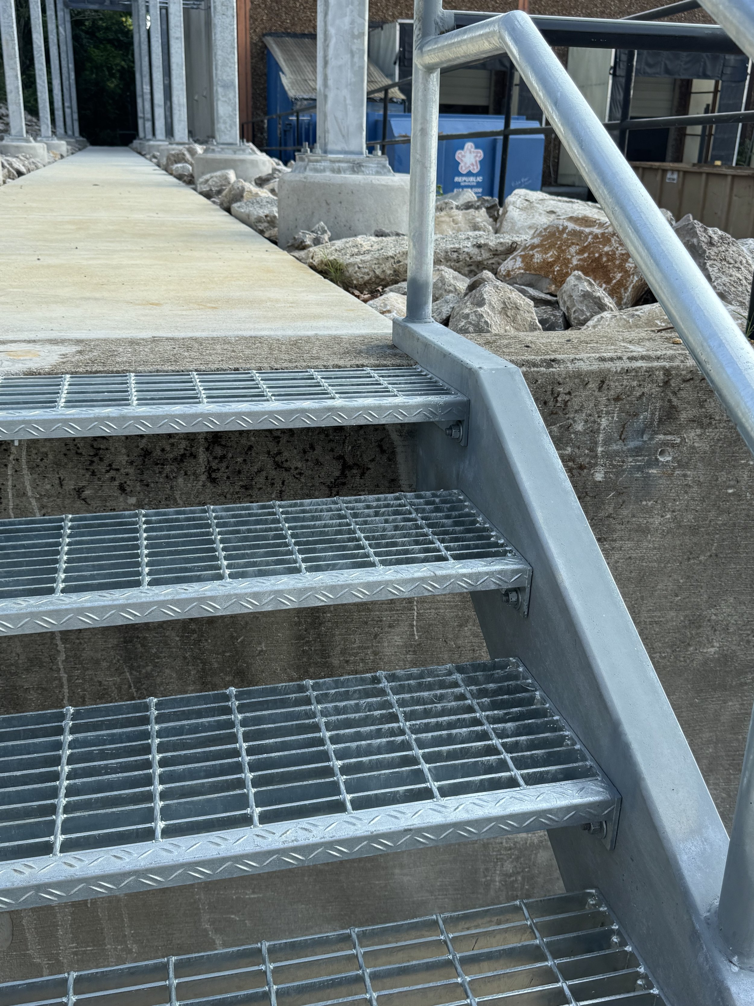 Metal outdoor stairs with four steps, metal railing on one side, concrete and rocks nearby, with construction materials and a blue container in the background.