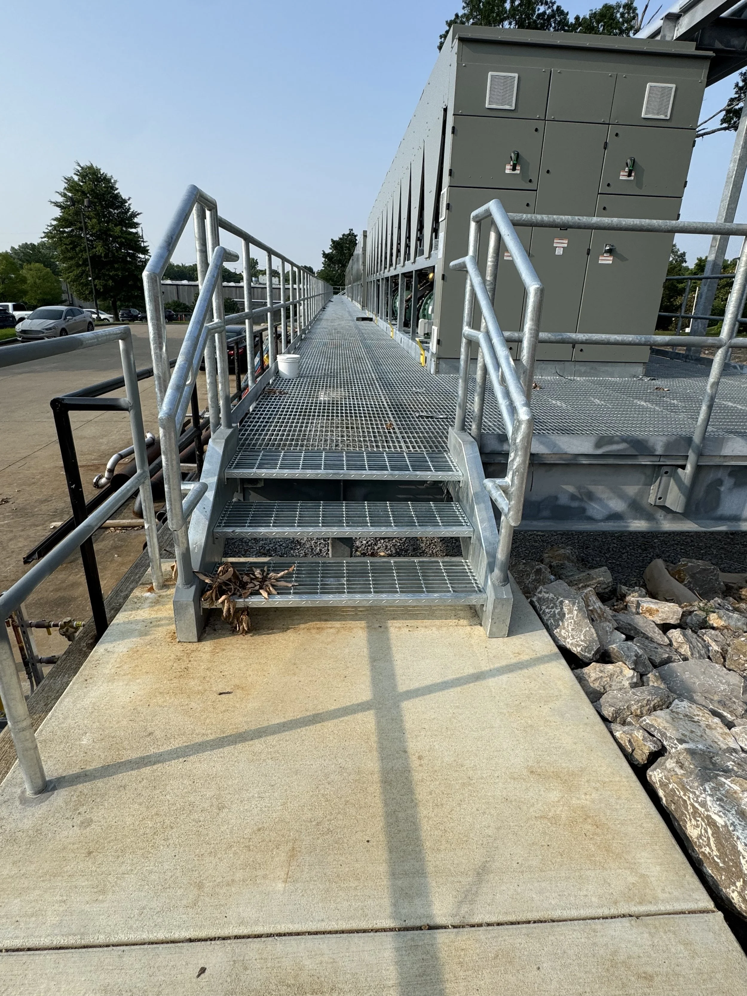 Metal walkway with stairs leading to a large electrical transformer or substation on an outdoor platform. Parking lot with cars and trees in the background, clear blue sky.