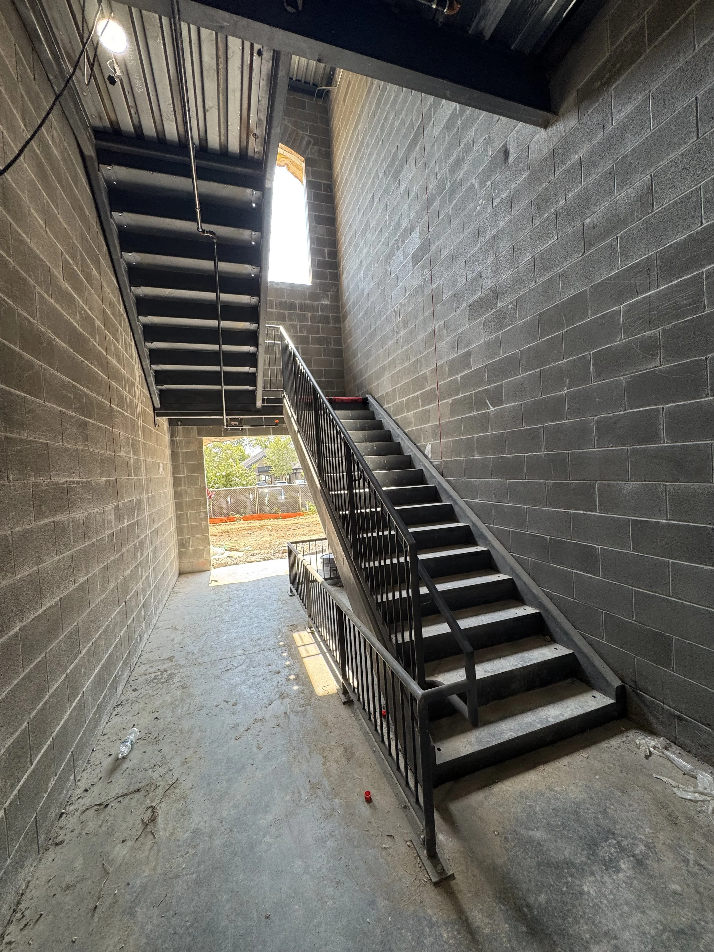 Industrial staircase inside a building with grey concrete walls and an open outdoor space visible at the top and bottom of the stairs.