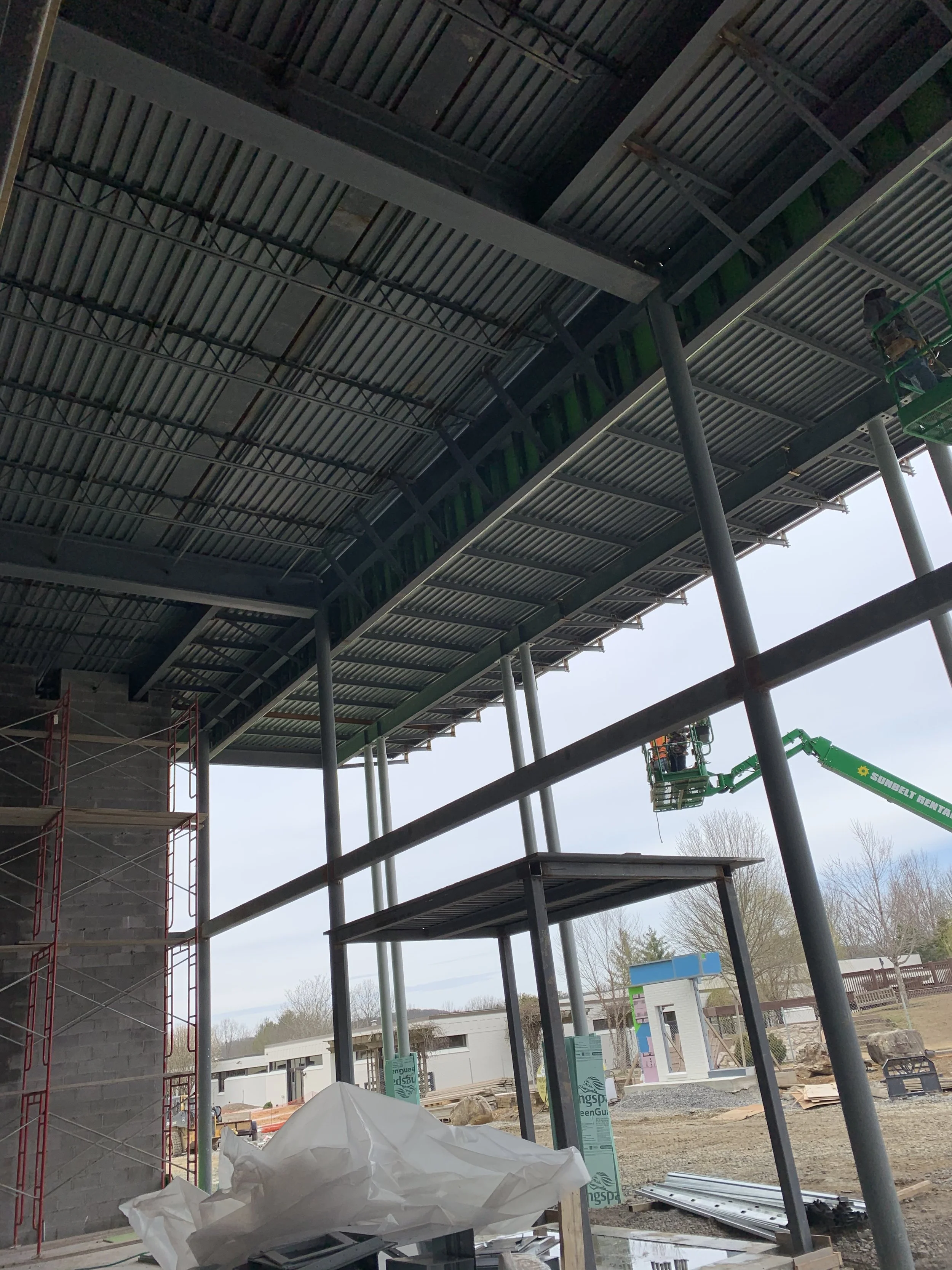 Construction site with workers in lift working on the underside of an elevated structure, with scaffolding to the left and a partially built wall, outdoor area with dirt and construction materials.