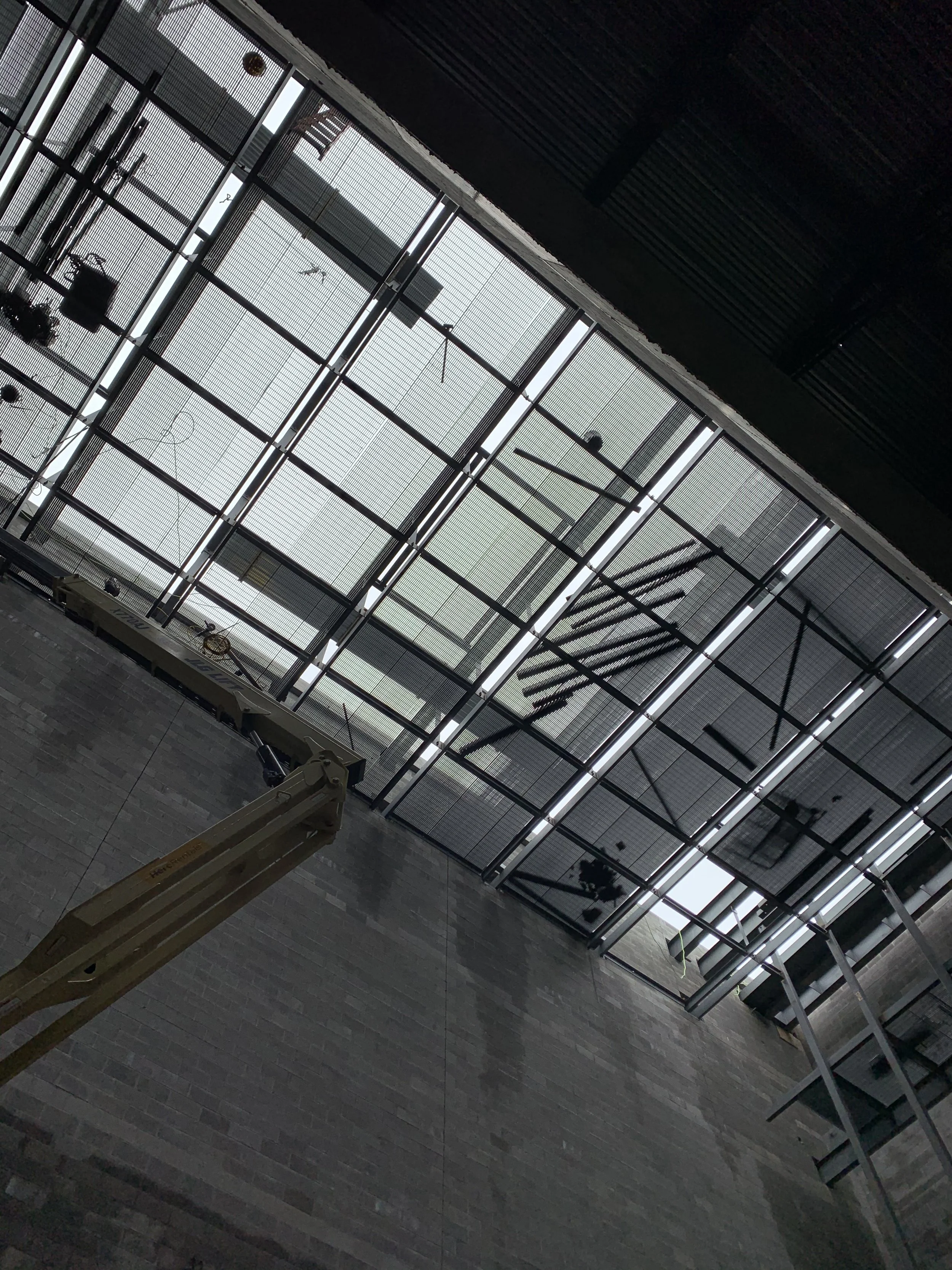 Looking up at a glass ceiling with grid pattern, metal beams, and some construction equipment and tools hanging or placed inside the building.
