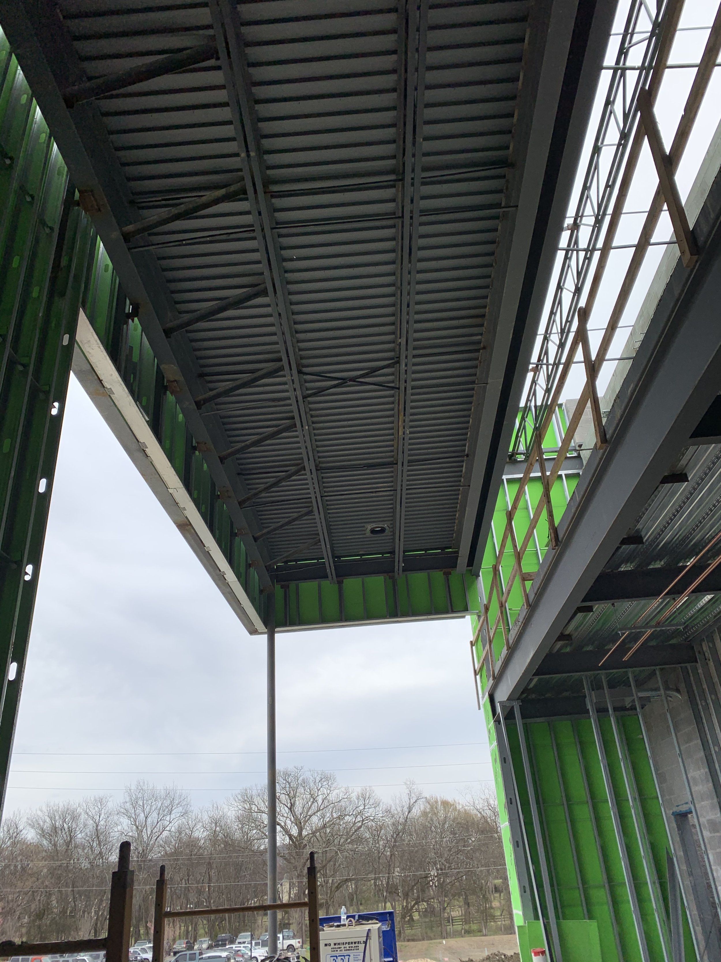 View of a construction site showing a partially built structure with steel framing, green sheathing, scaffolding, and an overhang roof, with trees and parked cars in the background.