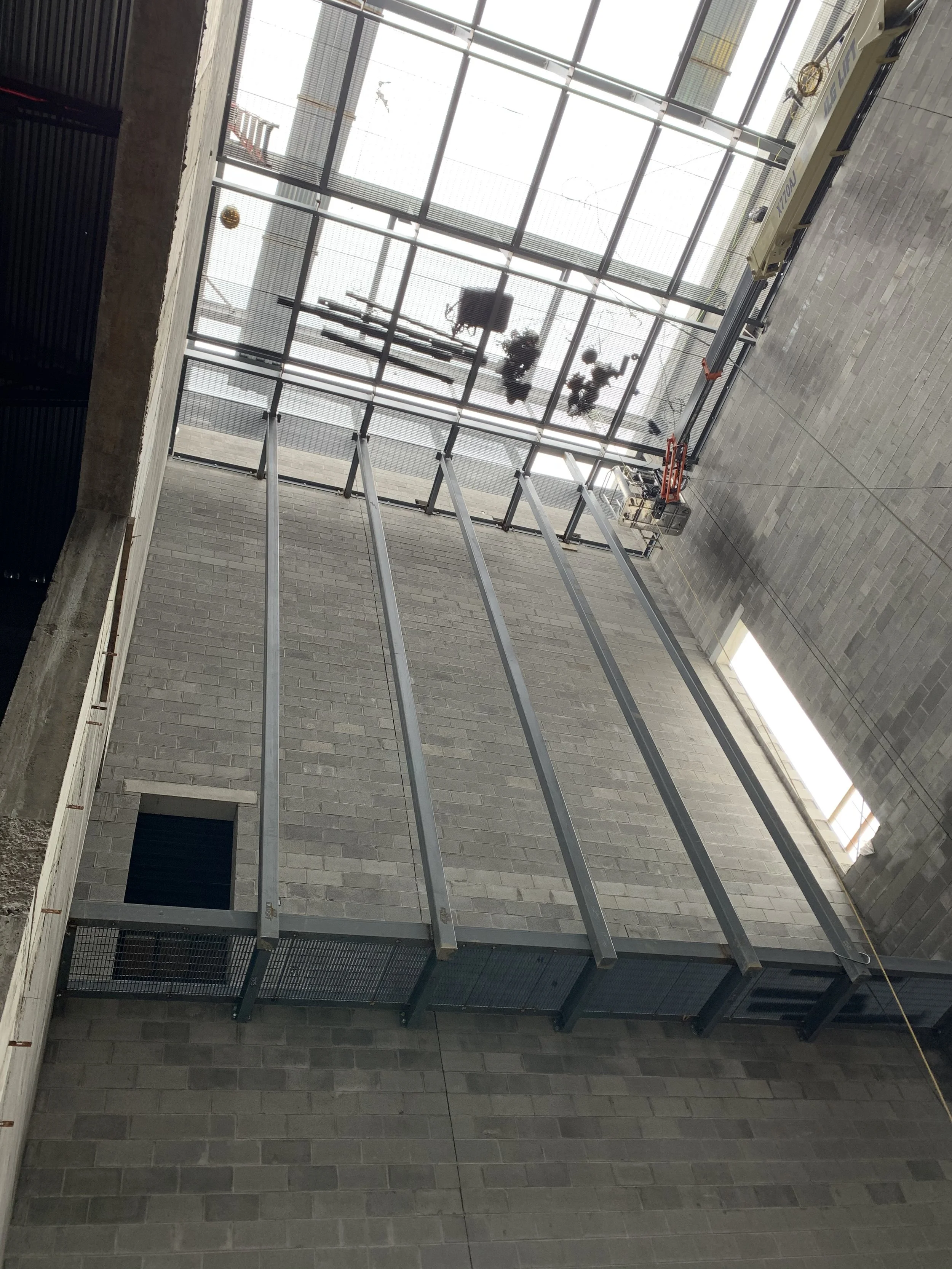 Interior view of a building under construction with steel beams, a gray brick wall, and large windows at the top letting in natural light.