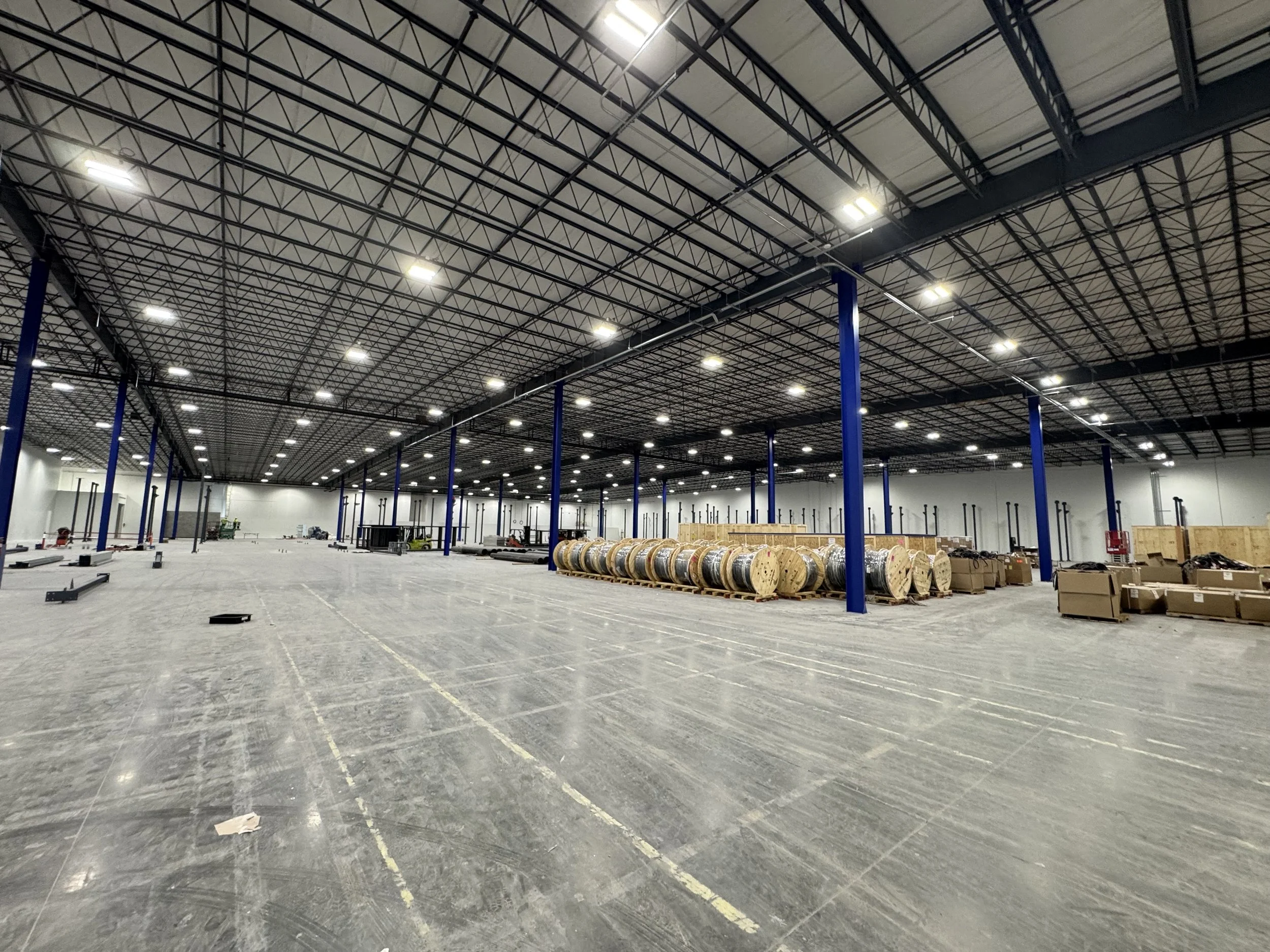 Inside a large, empty warehouse with a high ceiling, exposed metal beams, and bright overhead lighting. There are blue support columns, large rolls of cable or pipe, and stacks of wooden pallets and boxes along the sides.