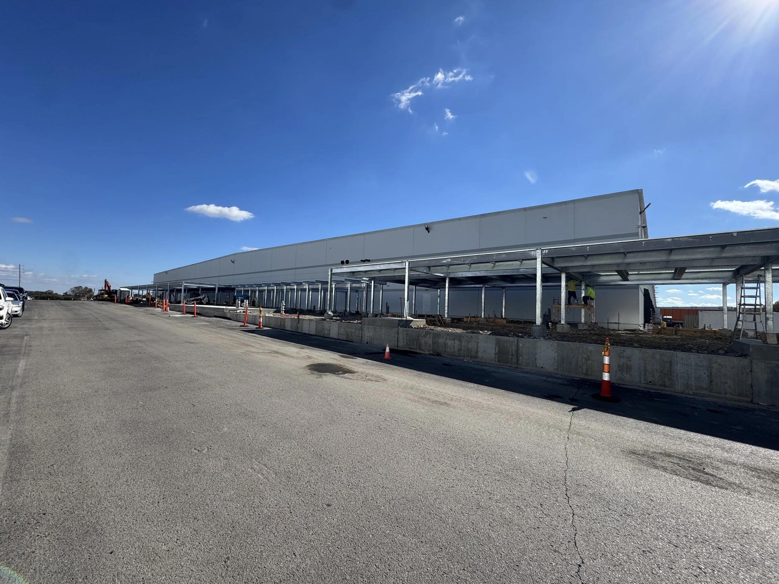 Construction site of a large commercial building with a white exterior and metal framework, partially covered walkway, orange cones, and construction workers in safety vests, under a clear blue sky.