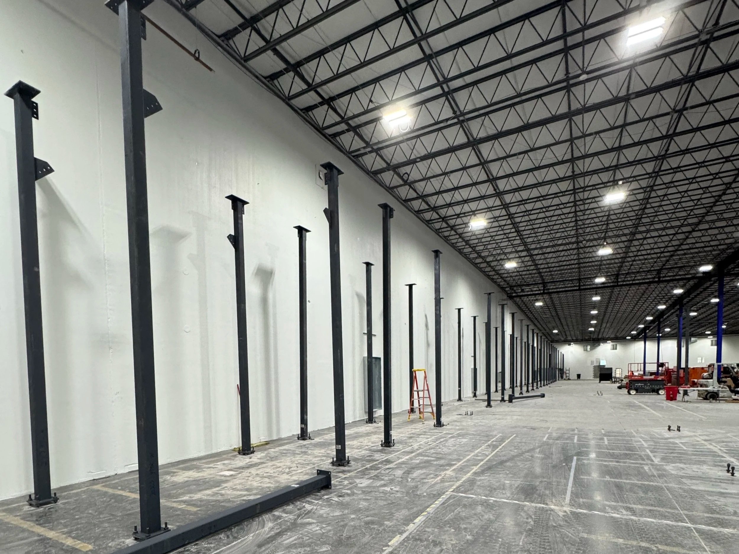 Inside a large warehouse or gymnasium under construction, with tall black metal support beams and a high ceiling, some construction equipment and scaffolding visible.