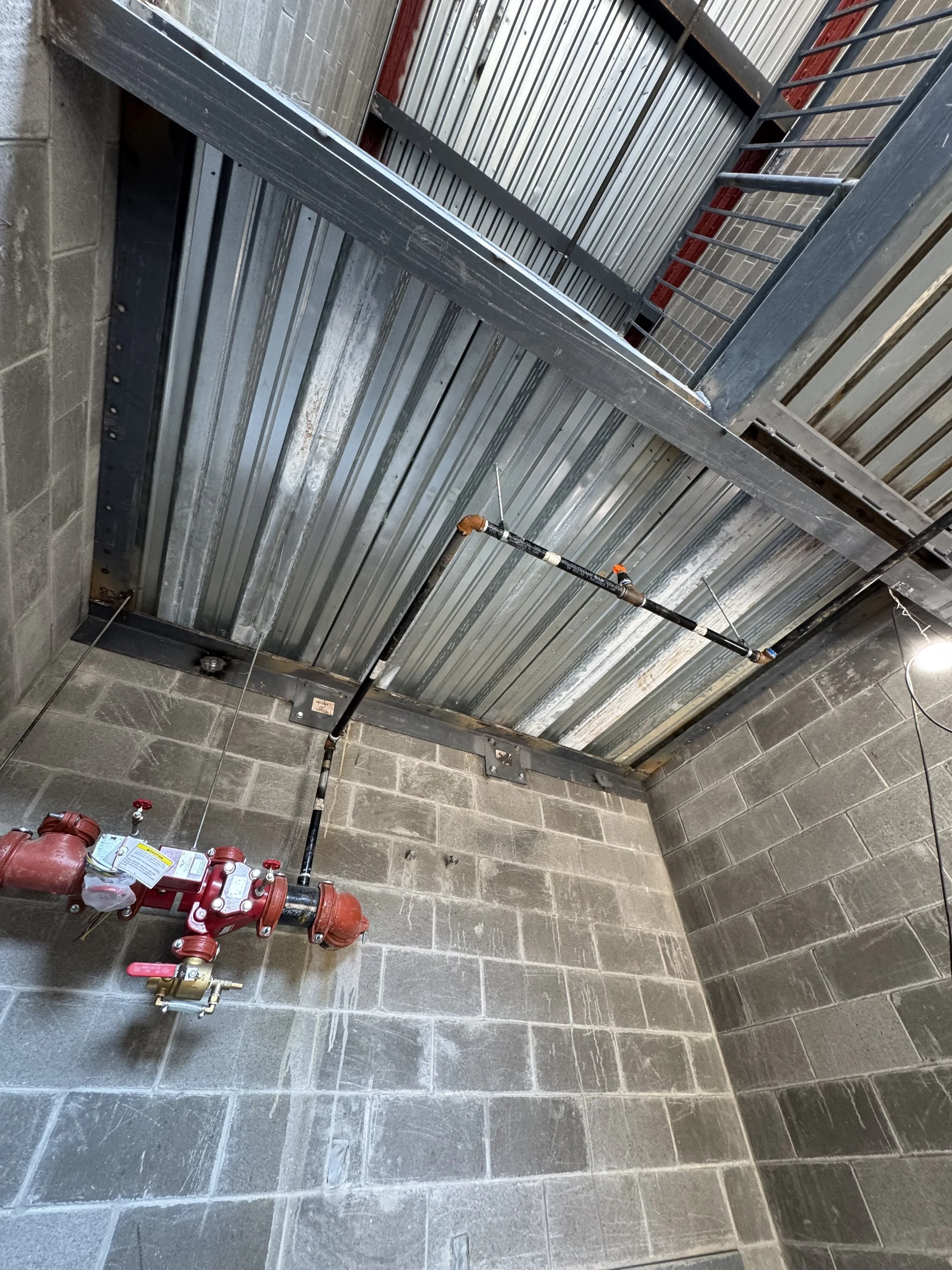 The image shows an industrial or commercial setting with a metal ceiling and a concrete block wall. There is a red fire valve connected to piping near the wall. A metal pipe extends from the valve and runs horizontally, supporting a long inspection r