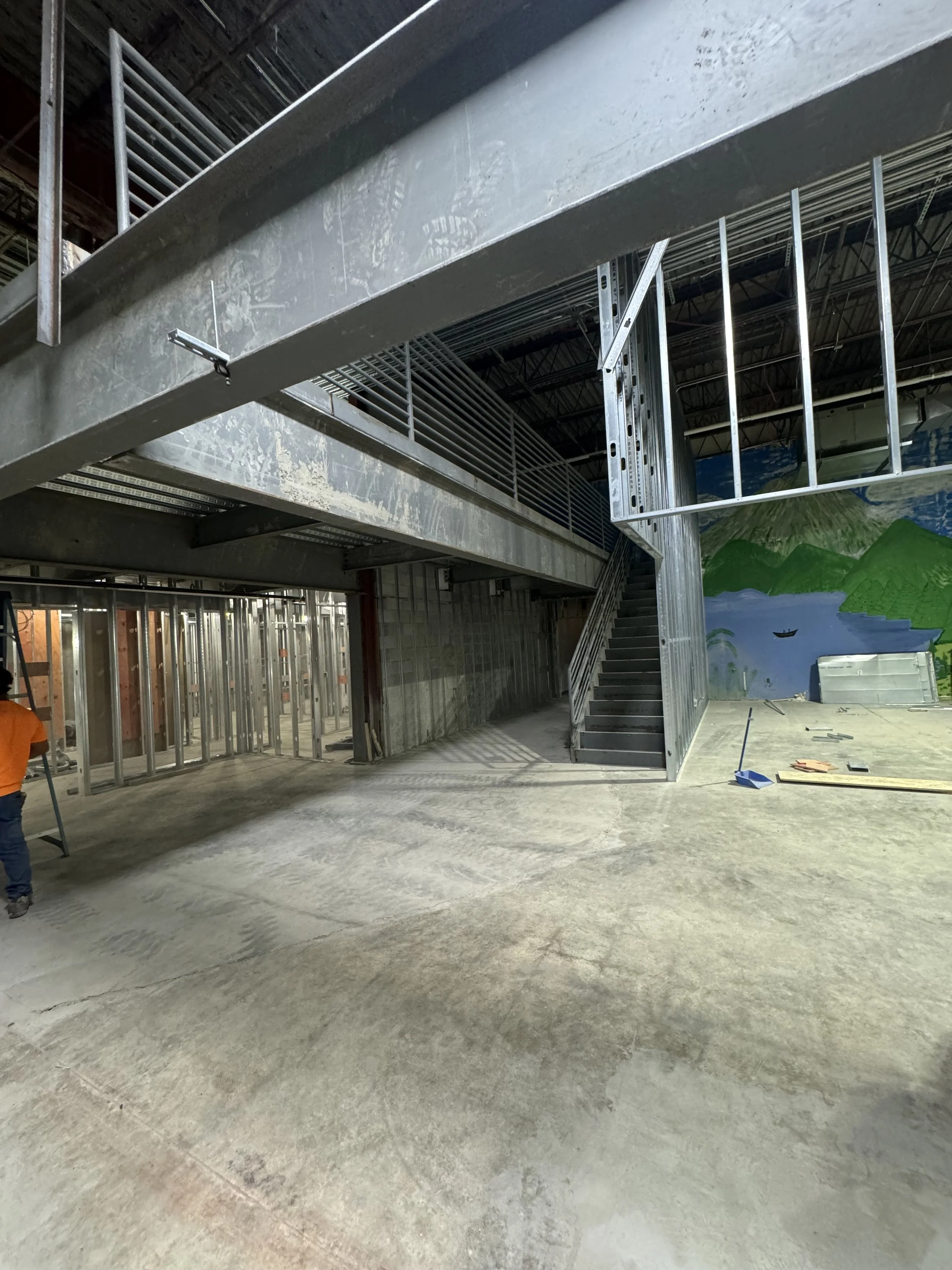 Interior of a building under construction with visible steel framing, stairs, and a partially painted mural on the wall featuring a landscape scene.