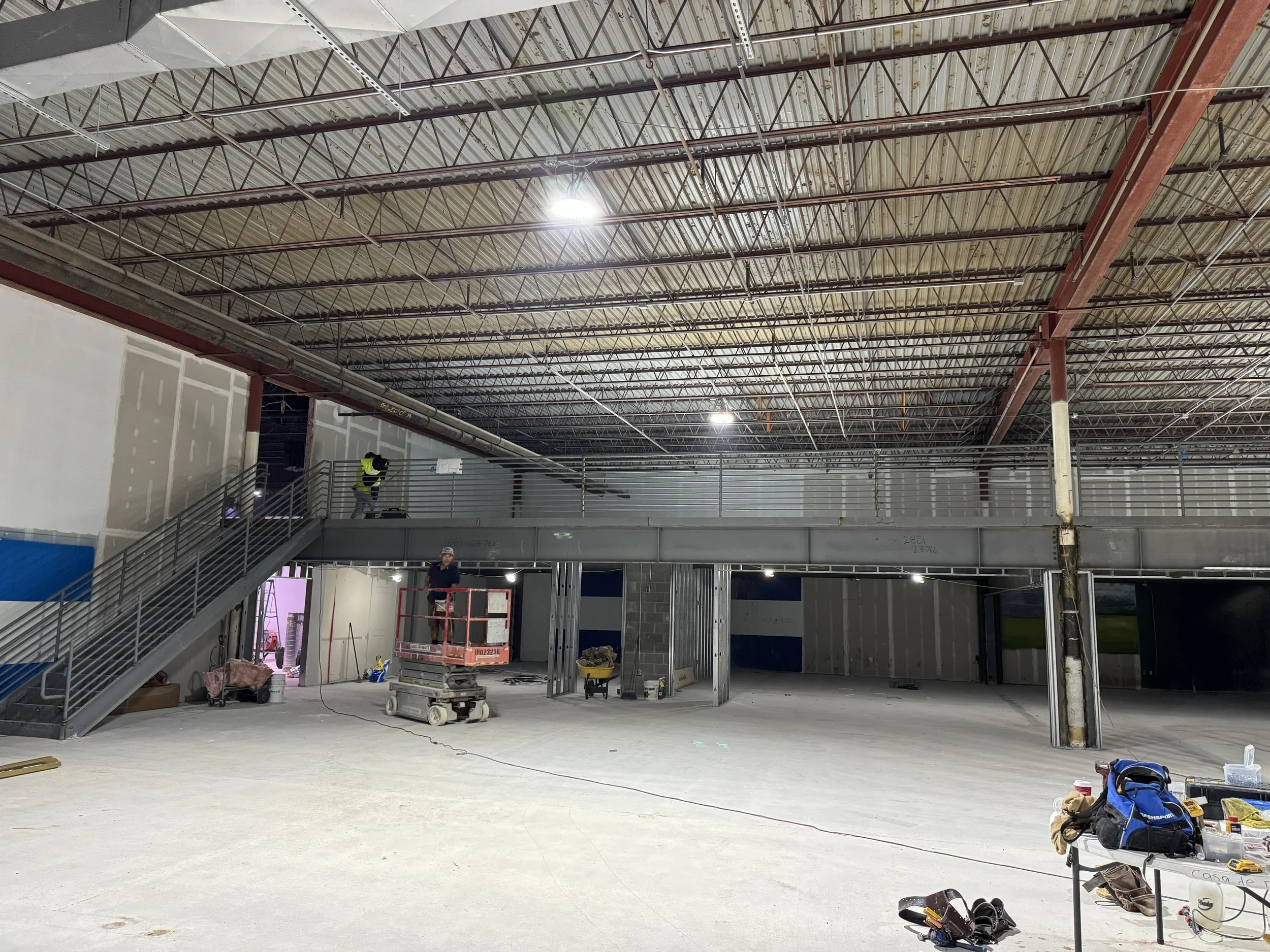Interior of a construction site with exposed metal framework, a staircase on the left, and workers installing a mezzanine level. Construction tools and supplies are scattered on the floor and a table in the foreground.