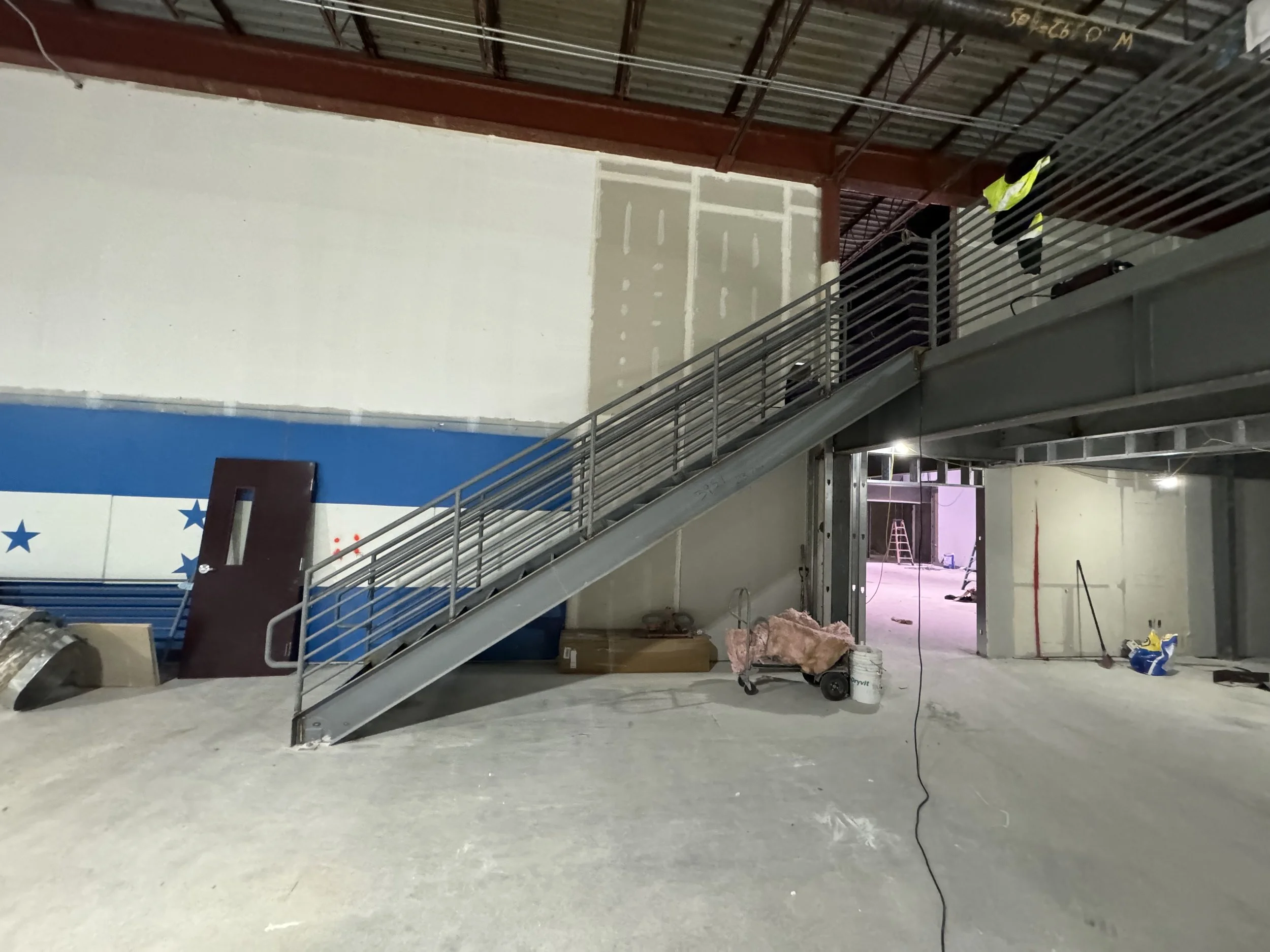 Construction site with steel staircase leading to an upper level, unfinished walls with drywall, construction tools, and equipment scattered on the floor.