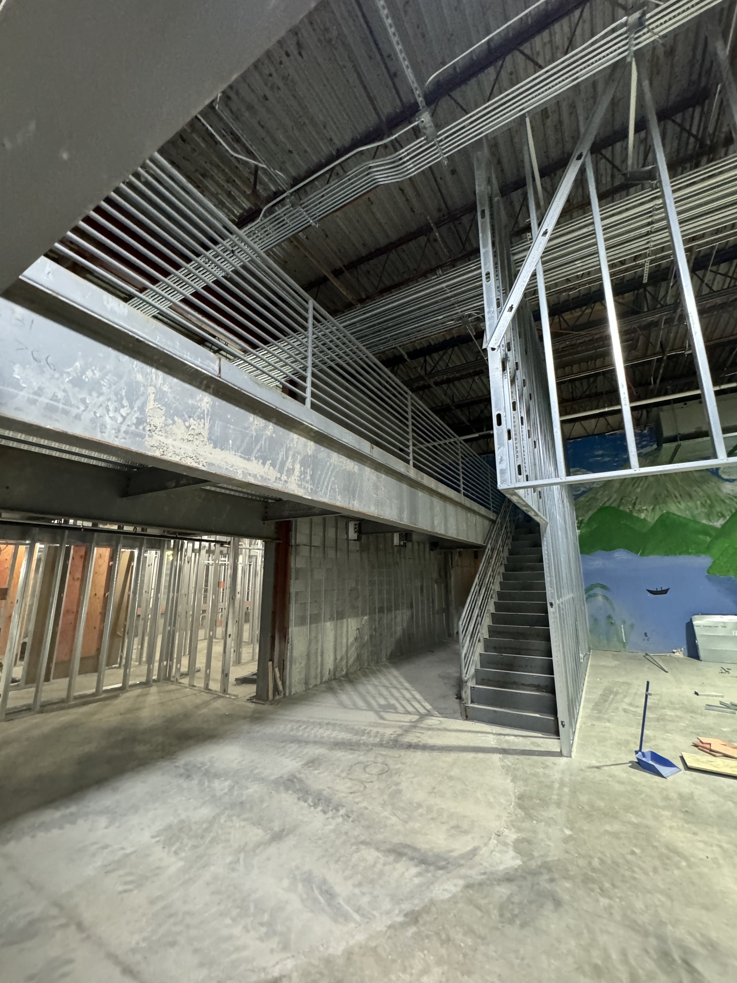 Interior of a building under construction with exposed metal framing, stairs, and a painted mural of mountains and water on a wall.