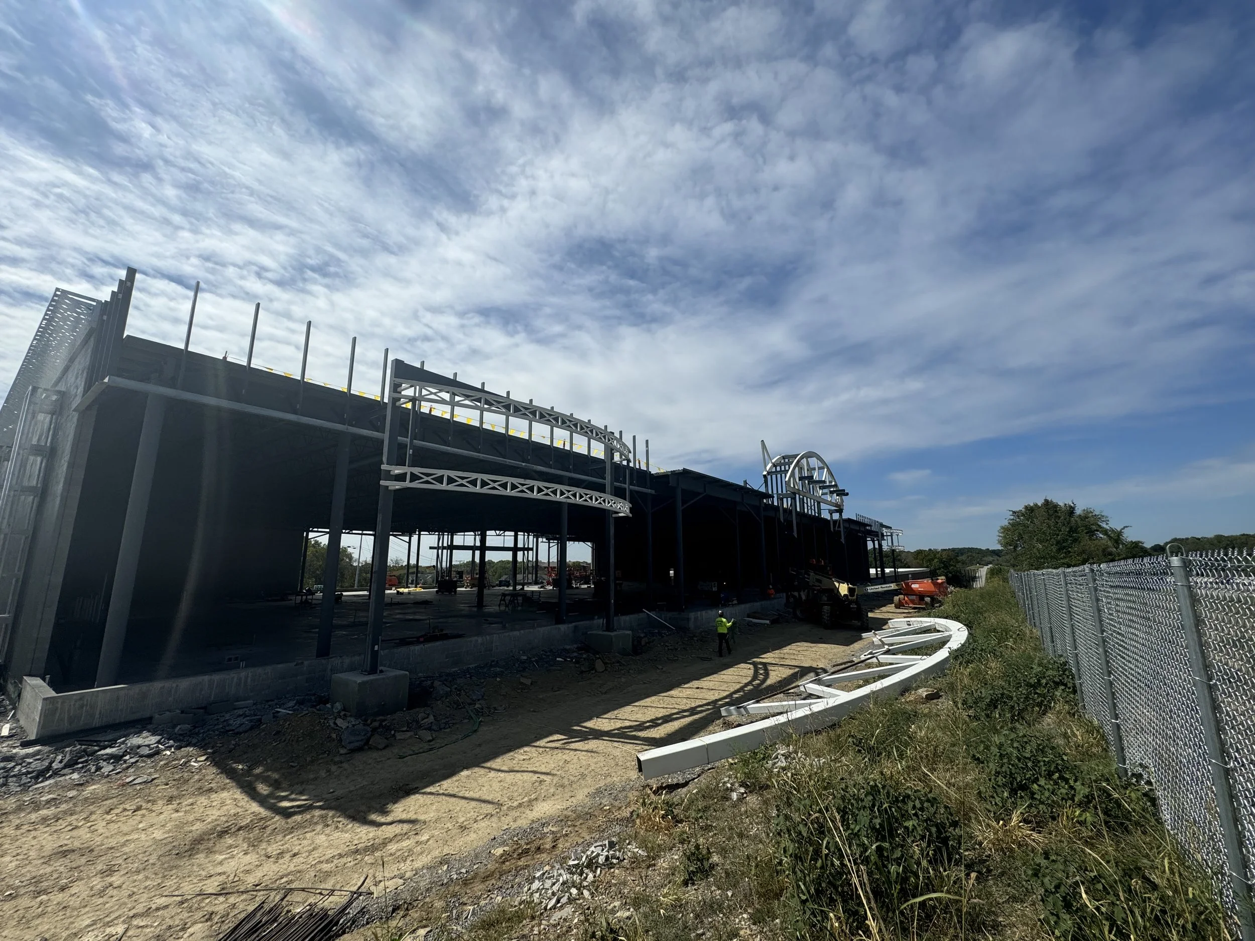 Under-construction building with metal framework, construction equipment, and workers, next to a chain-link fence under a partly cloudy sky.
