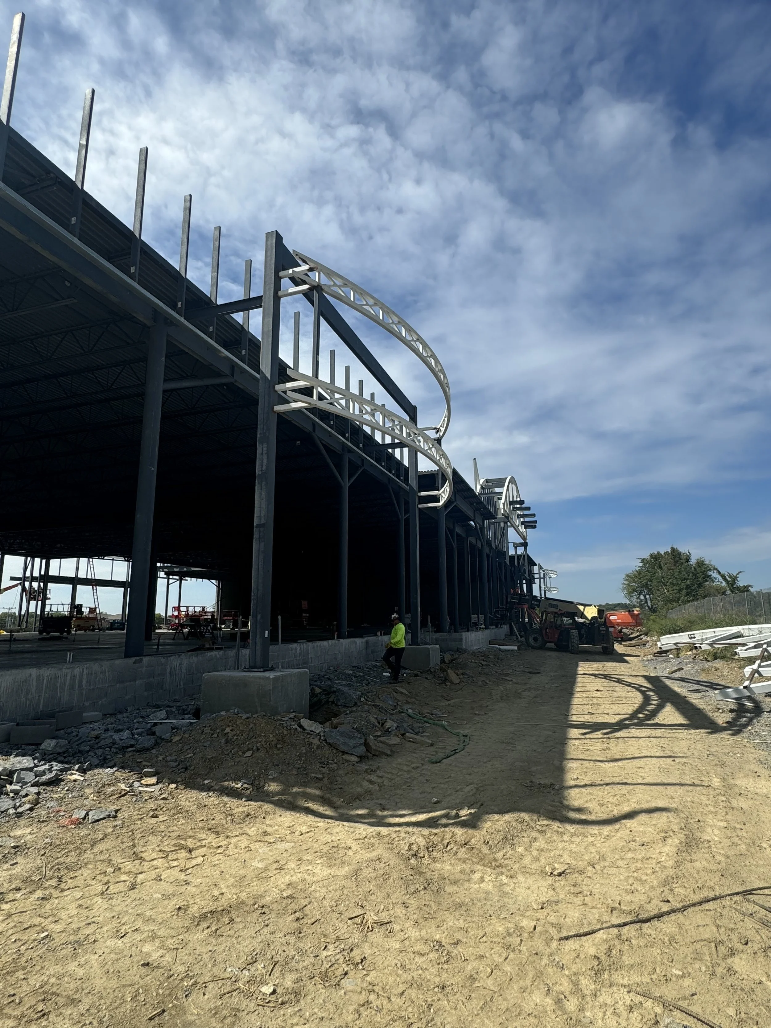 Construction site with a building framework and a dirt pathway, heavy machinery, and construction workers under a partly cloudy sky.