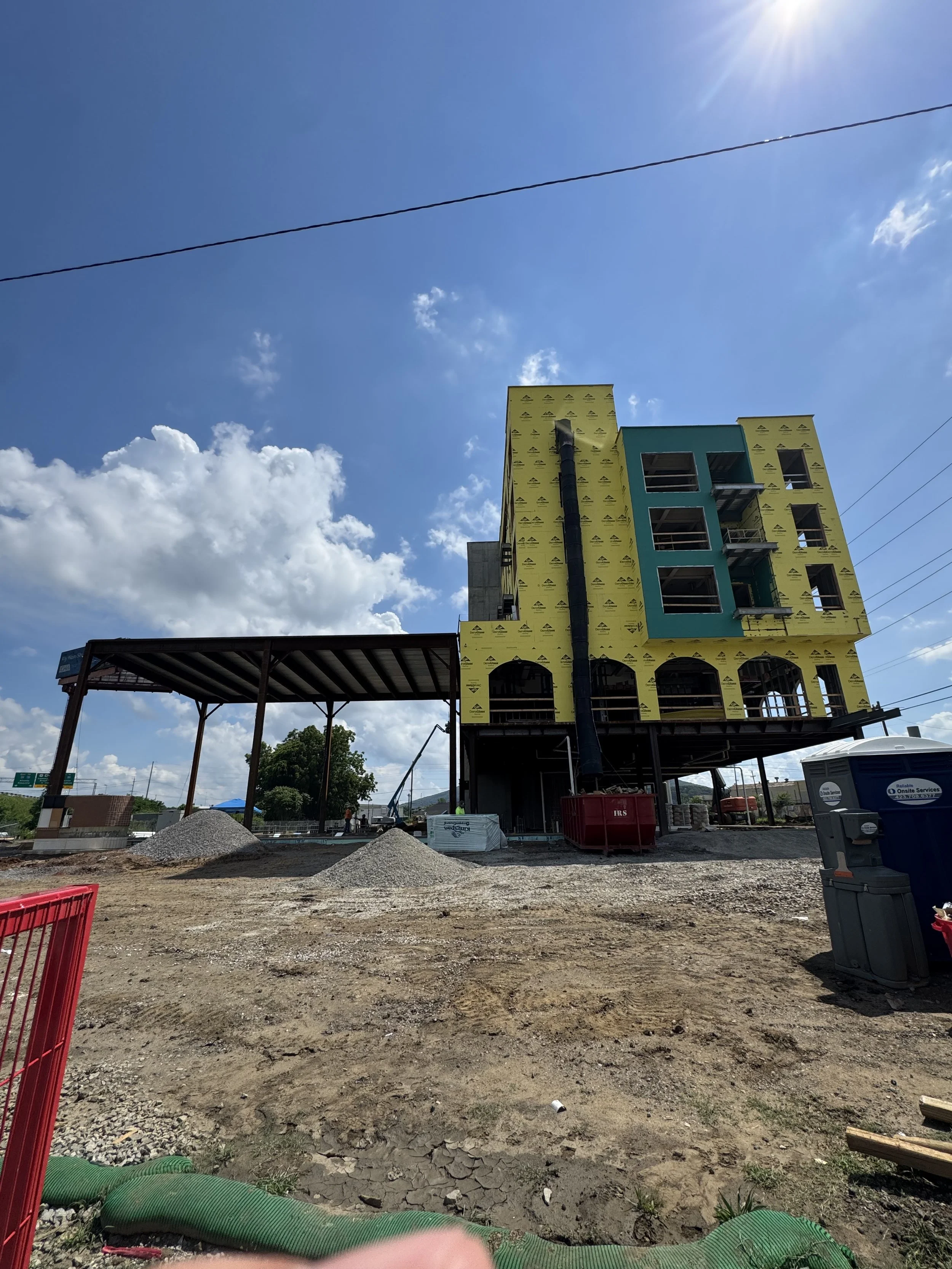 Under-construction multi-story building with yellow and teal facade, construction materials and equipment in foreground, construction site ground, clouds and blue sky overhead.