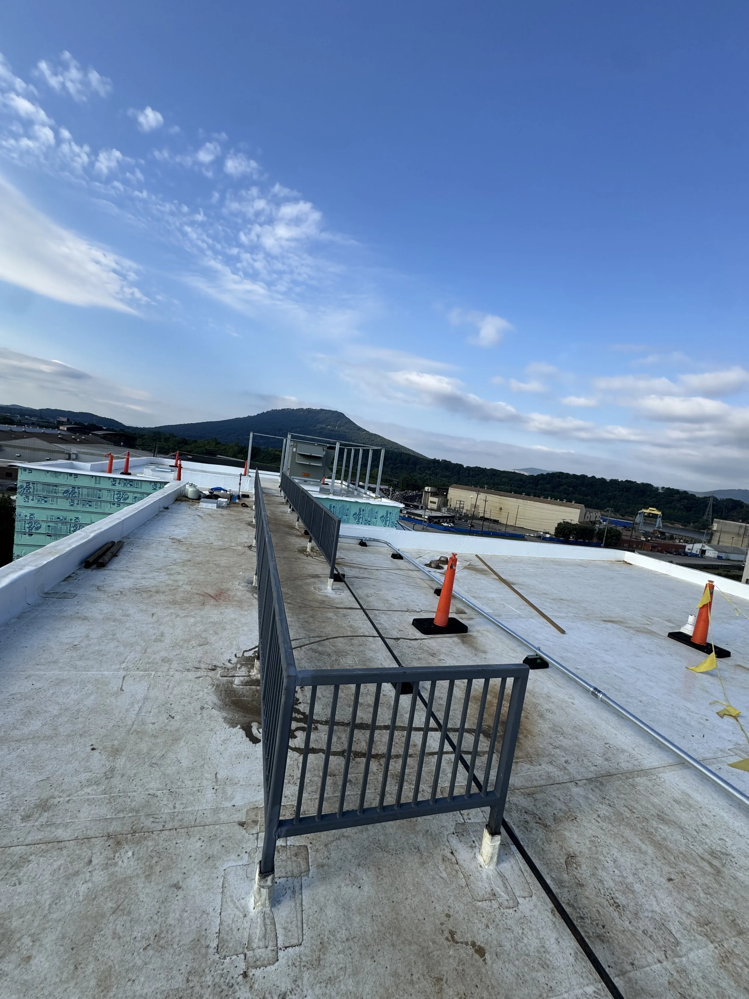 Construction site on a rooftop with safety barriers, orange cones, and construction materials, with a mountain and blue sky in the background.