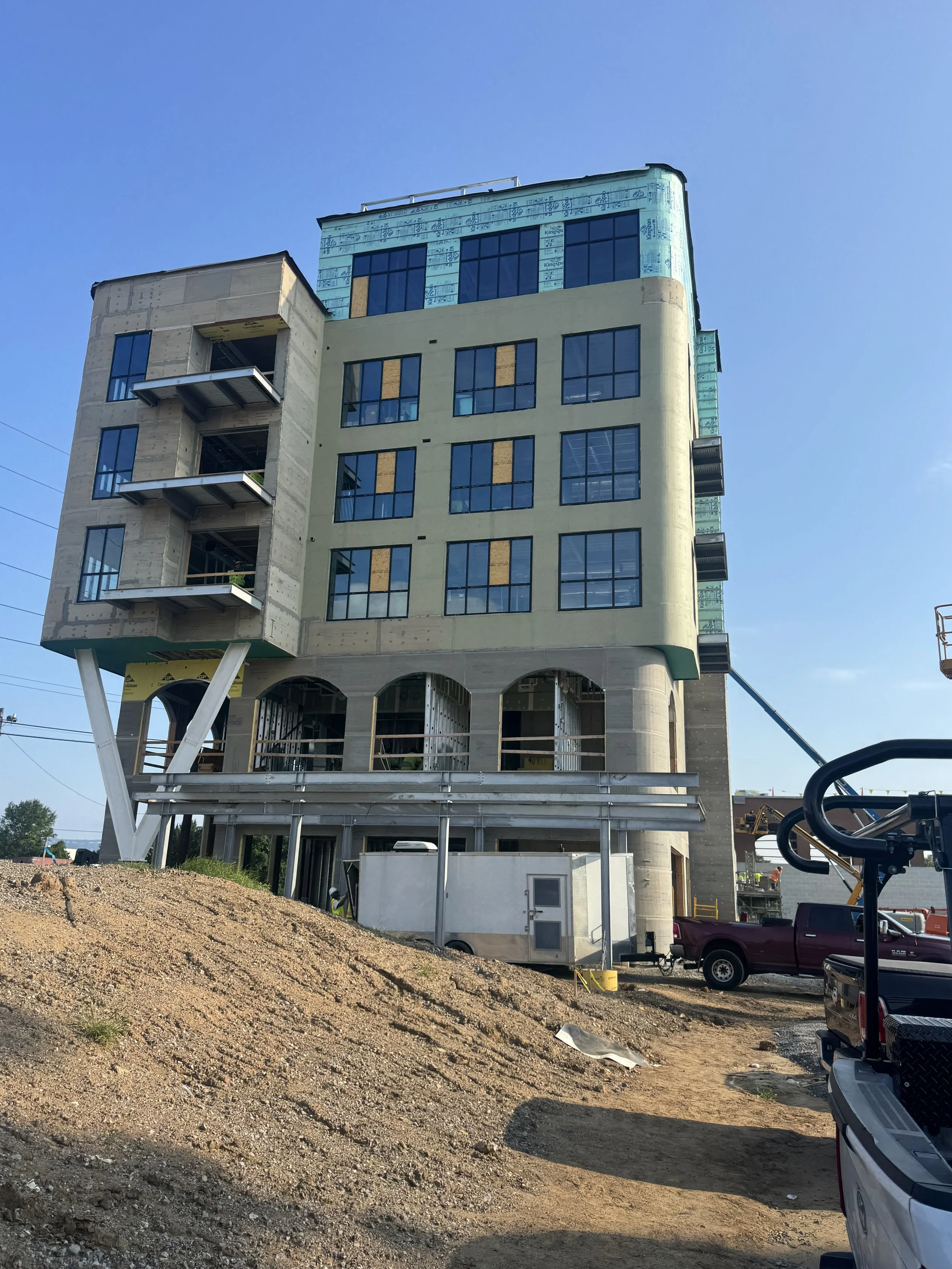 Under construction multi-story building with modern architectural design, exposed concrete, large windows, and construction equipment nearby, with a dirt ground in the foreground.