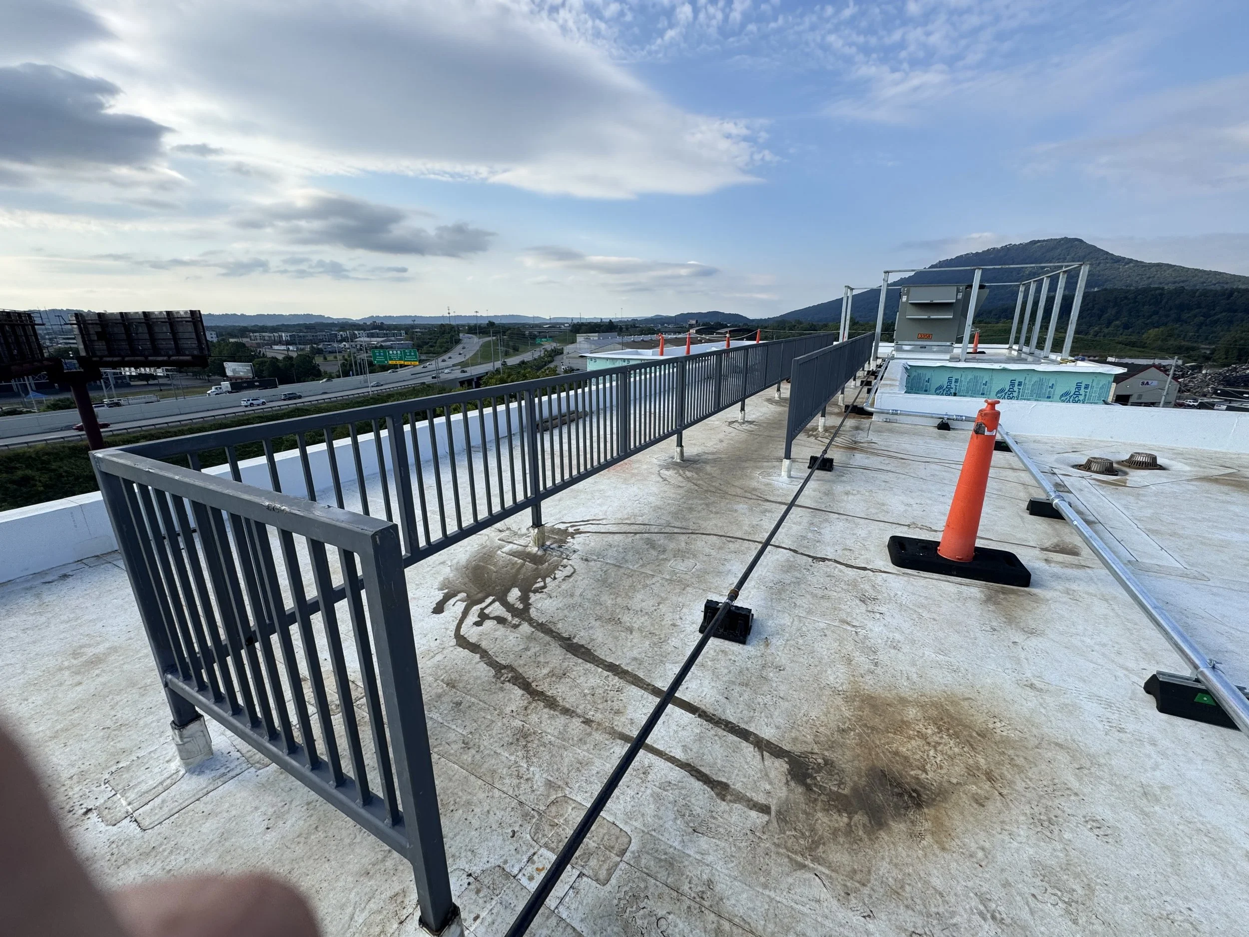 On a rooftop, a safety railing is partially installed, with construction cones, equipment, and tools visible on the roof, overlooking a highway and distant mountains under a partly cloudy sky.