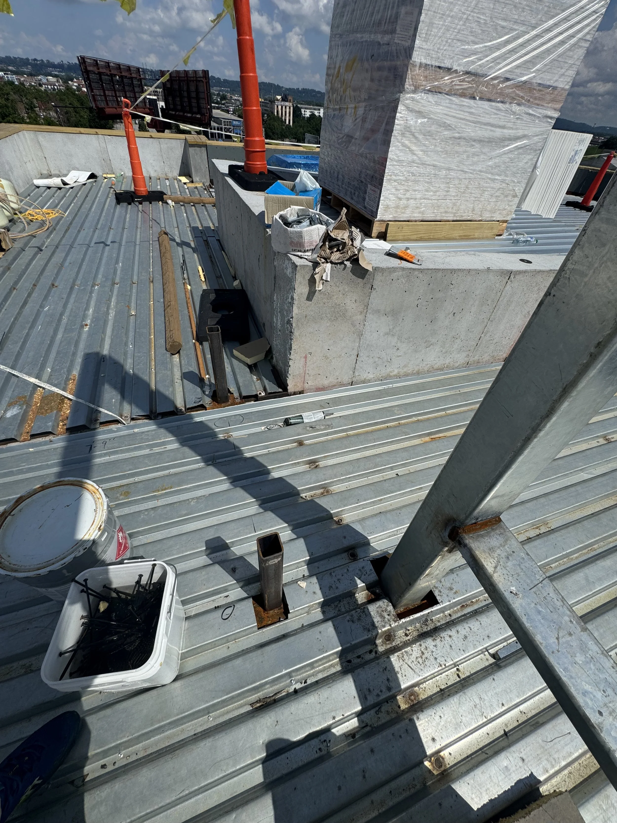 View of a rooftop under construction with tools, building materials, and safety equipment, including a concrete chimney and metal railing, under partly cloudy sky.