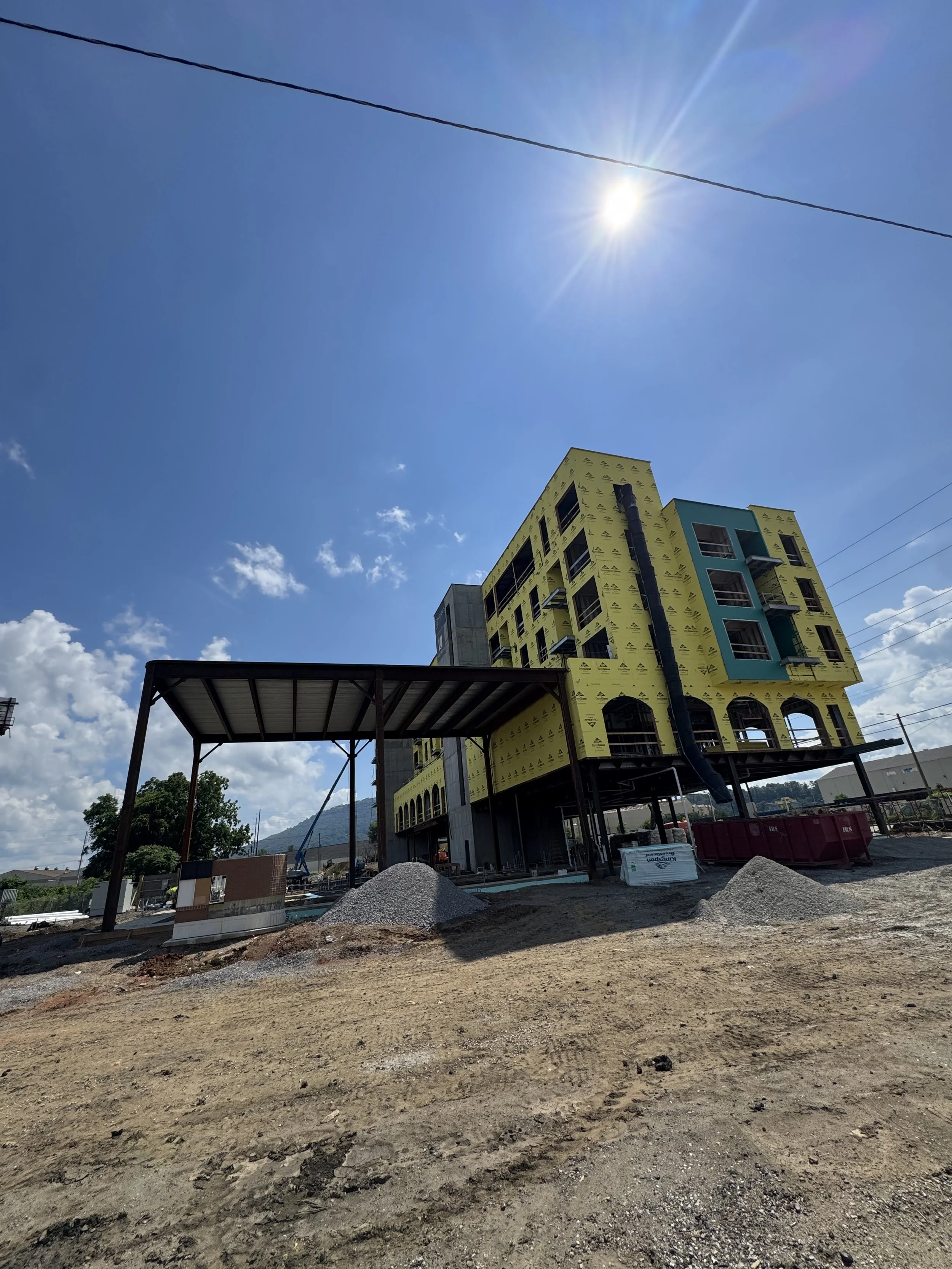 Under-construction building with yellow wrapping, black drainpipe, and attached slide, on a dirt lot with piles of gravel, under a sunny sky.